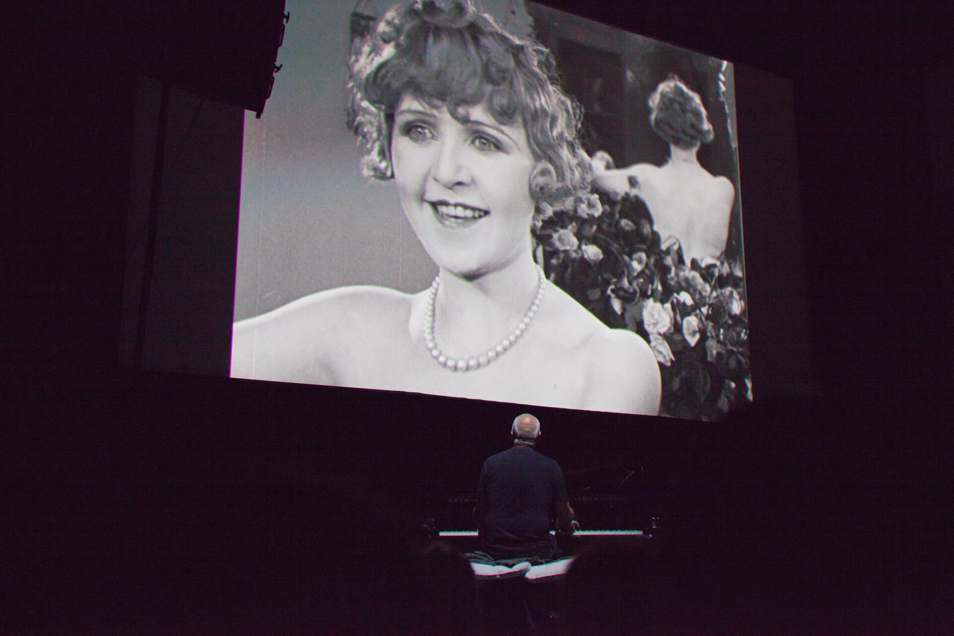 Zdjęcie ukazuje duży ekran, na którym widać młodą kobietę z dawnych filmów. Pod ekranem siedzi mężczyzna i patrzy na ekran. 