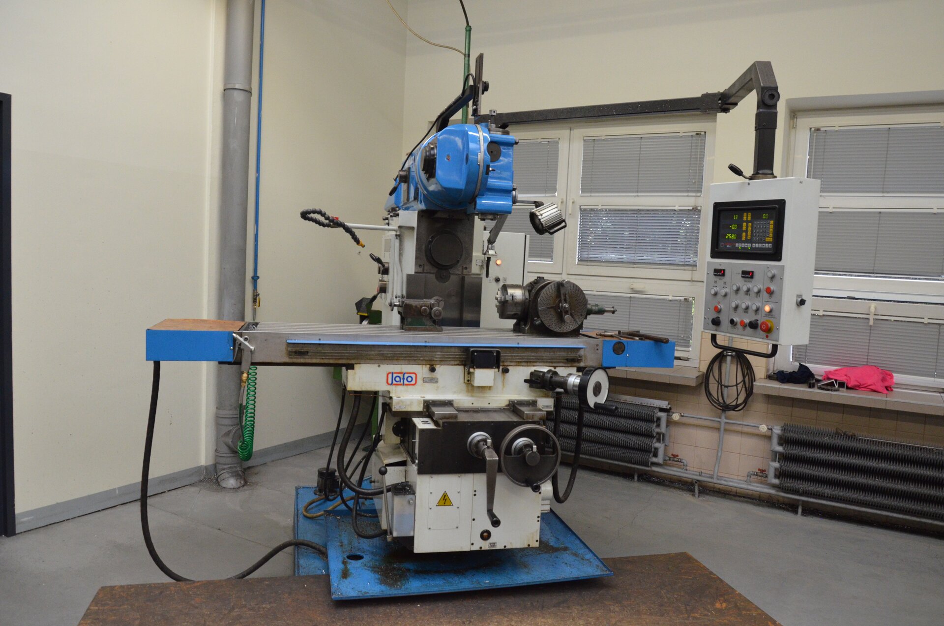 Ilustracja przedstawia frezarkę koloru niebieskiego. Frezarka znajduje się w warsztacie, jest zbudowana z żeliwnego korpusu, na którym pionowo umieszczono  napędzane wrzeciono, stół frezarki posiada posuw poprzeczny, wzdłużny i w płaszczyźnie pionowej. 