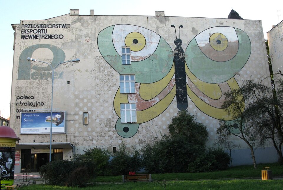 Zdjęcie przedstawia ścianę budynku dawnego Pewexu z muralem w kształcie motyla. Po lewej stronie budynku widnieje napis „Przedsiębiorstwo eksportu wewnętrznego”. Budynek ma trzy okna. Motyl namalowany jest w kolorach - zielony, żółty, czerwony oraz czarny. Przed budynkiem ukazana jest ławka oraz żółty śmietnik.