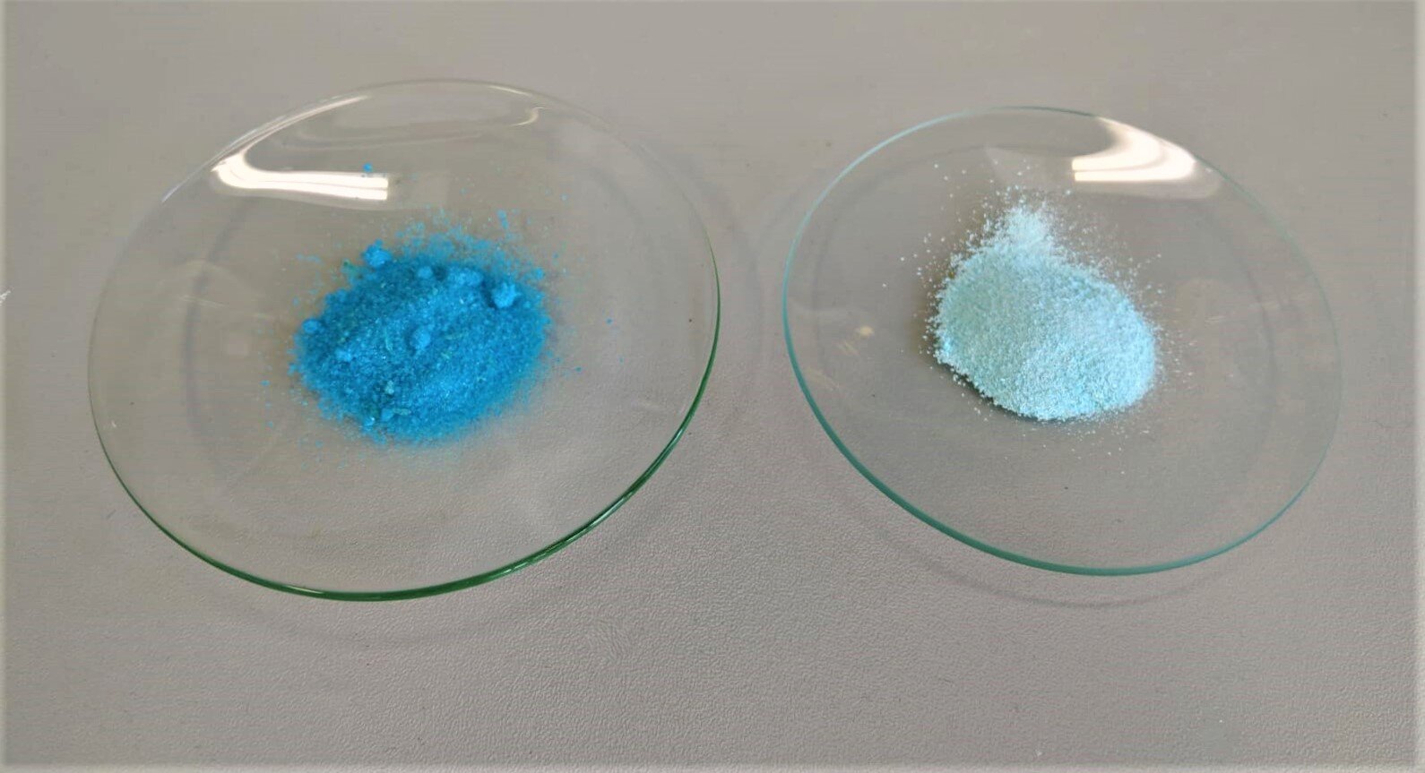 Zdjęcie przedstawia dwa szkiełka zegarkowe. Na jednym z nich jest pewna ilość niebieskiego proszku, na drugim jasnoniebieskiego.