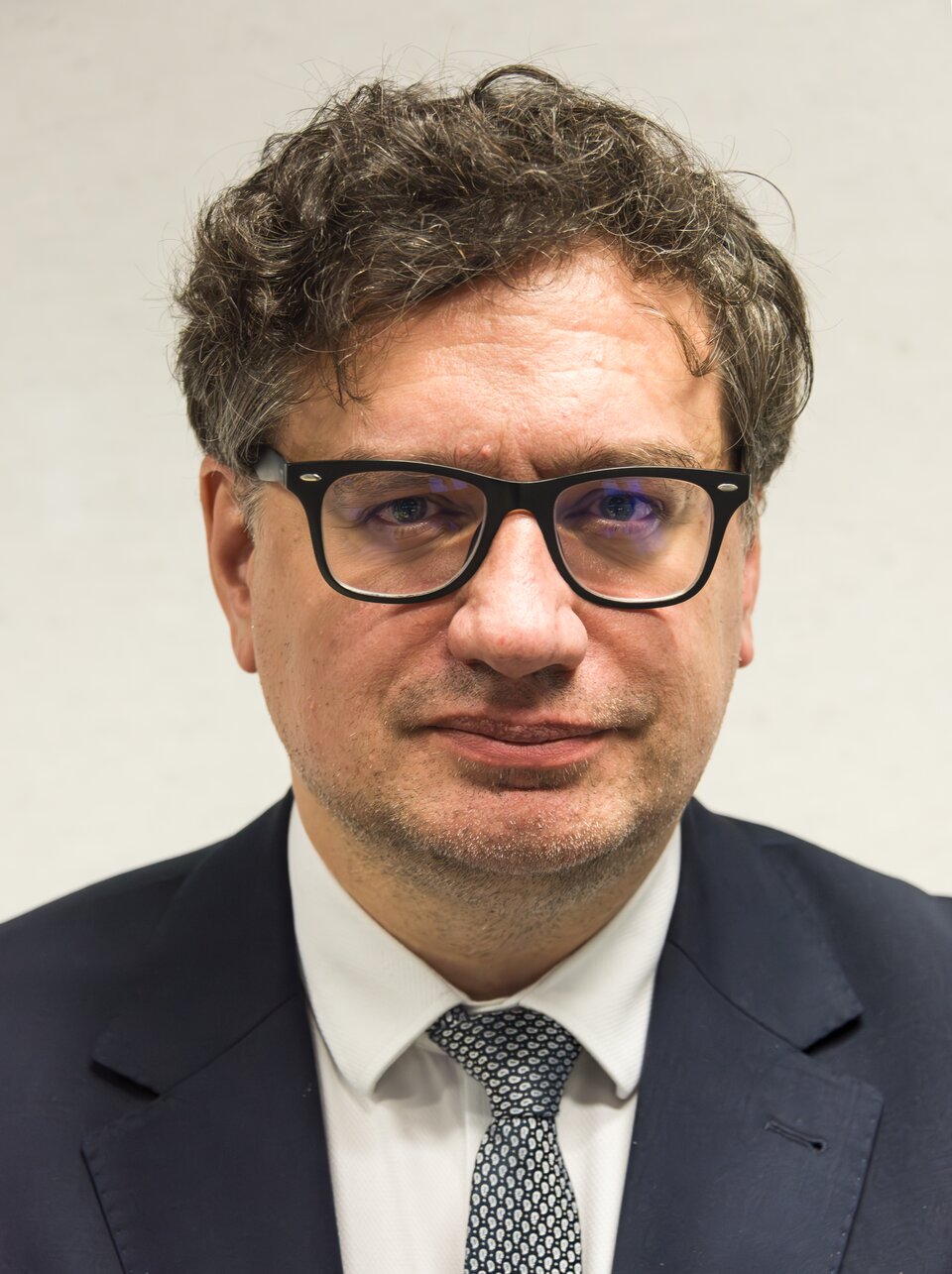 Portret mężczyzny w średnim wieku w okularach w grubych, czarnych oprawkach, ubranego w ciemną marynarkę, białą koszulę i wzorzysty krawat.