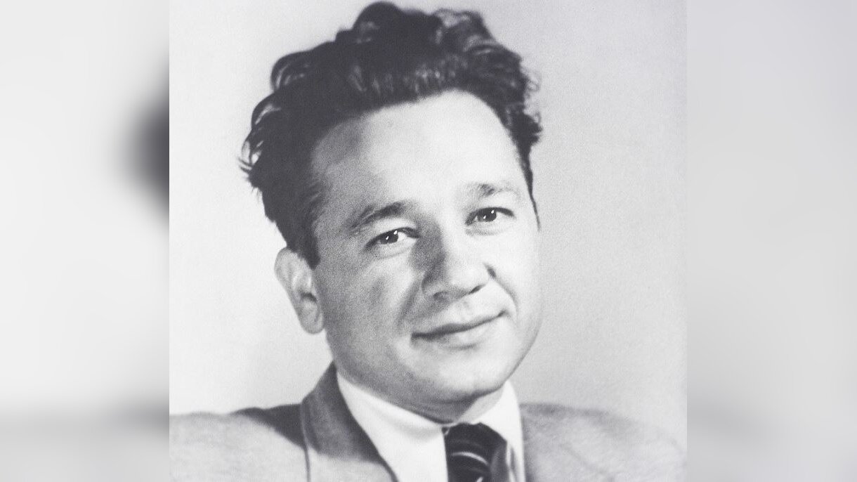 Zdjęcie przedstawia portret mężczyzny. Mężczyzna ma ciemne włosy zaczesane do tyłu, lekko się uśmiecha. Ubrany w garnitur, koszulę i krawat.