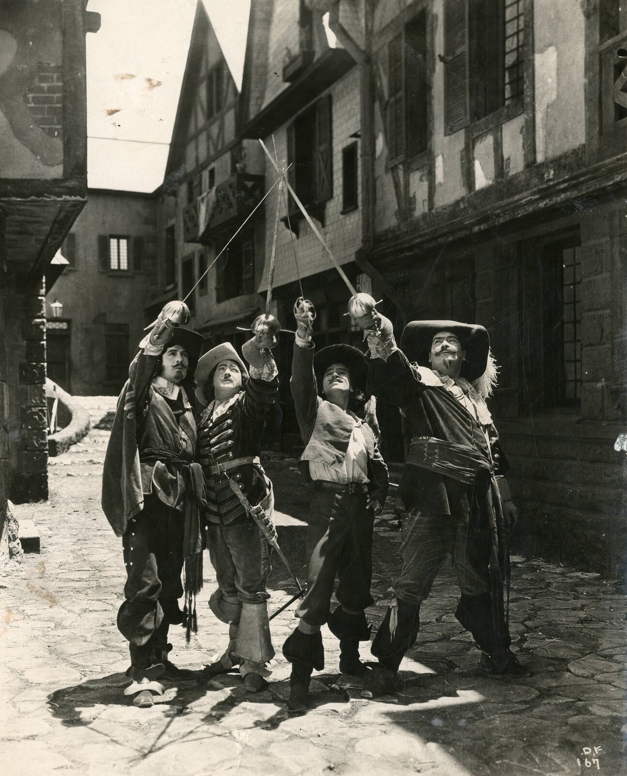 Zdjęcie przedstawia czterech mężczyzn stojących na tle starych budynków wykonanych z cegły i drewna. Są oni ubrani podobnie - każdy z nich ma na głowie kapelusz z piórem, a także koszule i bogato zdobione marynarki. Ich spodnie są bufiaste i włożone do wyższych butów skórzanych. Unoszą szable ku górze, złączając je czubkami. Zdjęcie jest czarno‑białe.