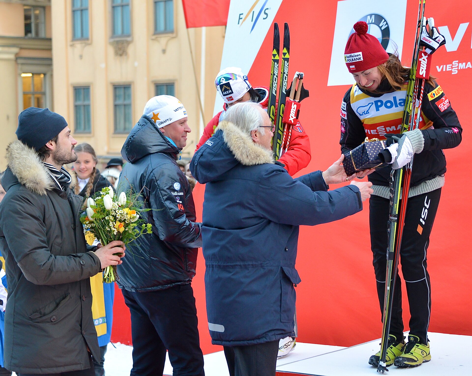 Zdjęcie przedstawia Justynę Kowalczyk na najwyższym stopniu podium.  Ubrana jest w strój sportowy, czarne dresy z koszulką reklamową sponsorów i czerwoną czapkę. W ręku trzyma narty. Trzech mężczyzn składa jej gratulacje. Jeden trzyma bukiet kwiatów, drugi wręcza jej pudełeczko z nagrodą. Justyna szeroko się uśmiecha.