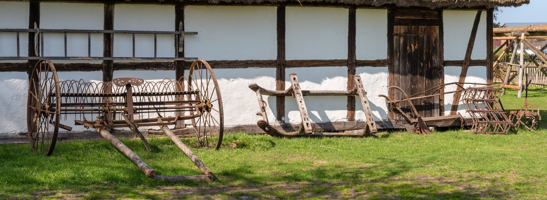 Kliknij, aby powiększyć. Zdjęcie przedstawia narzędzia rolnicze - pług, orkę, radło - stojące przy bocznej ścianie chaty. Budynek ma konstrukcję szkieletową złożoną z drewnianych belek wypełnionych murem, który jest bielony.