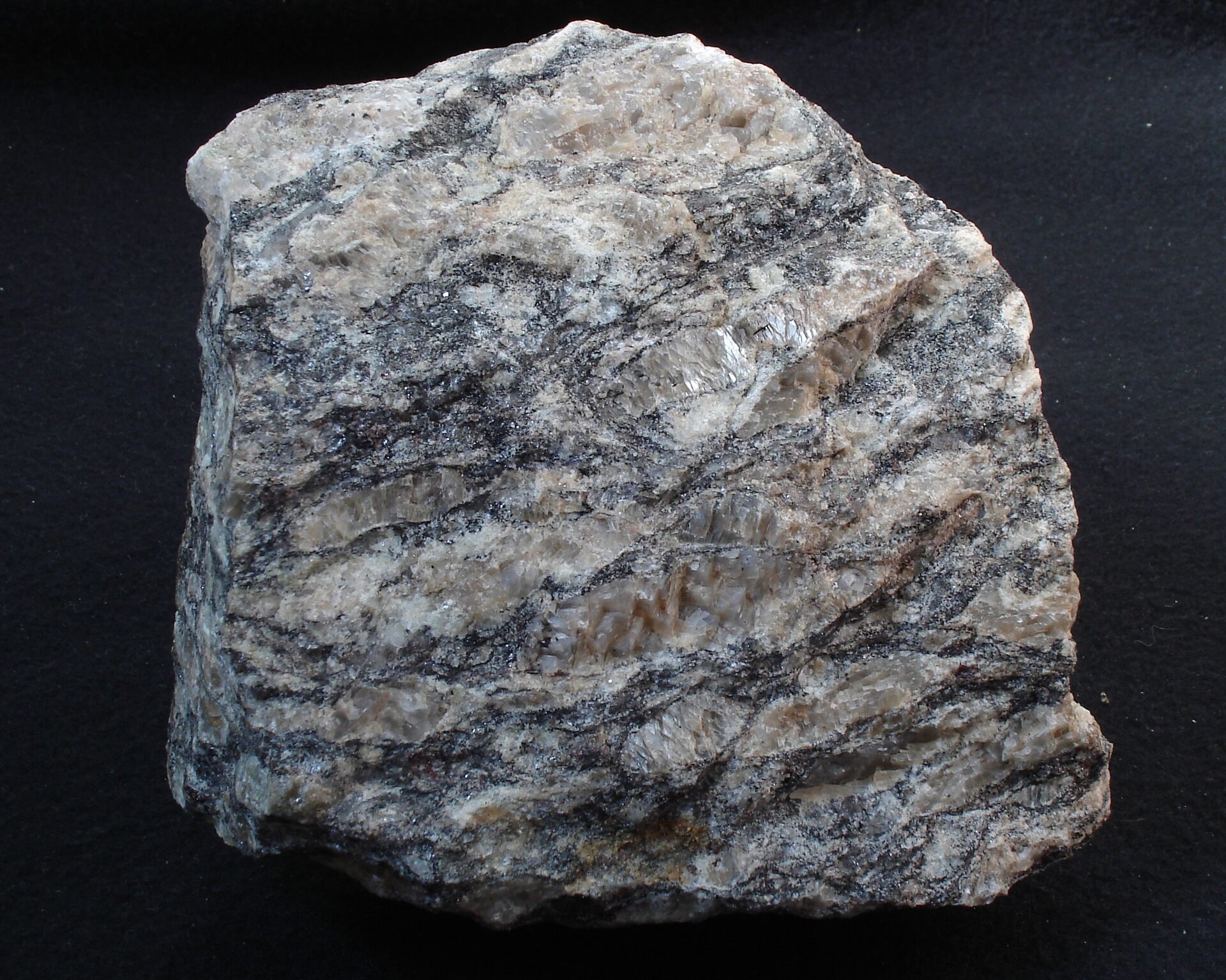 Na zdjęciu przedstawiony jest gnejs – barwa szara, warstwy kwarcowe jaśniejsze różowe, a łyszczykowe ciemniejsze srebrzyste i czarne. Równoległe ułożenie minerałów, tekstura kierunkowa. Fragment skały wielkości pięści.