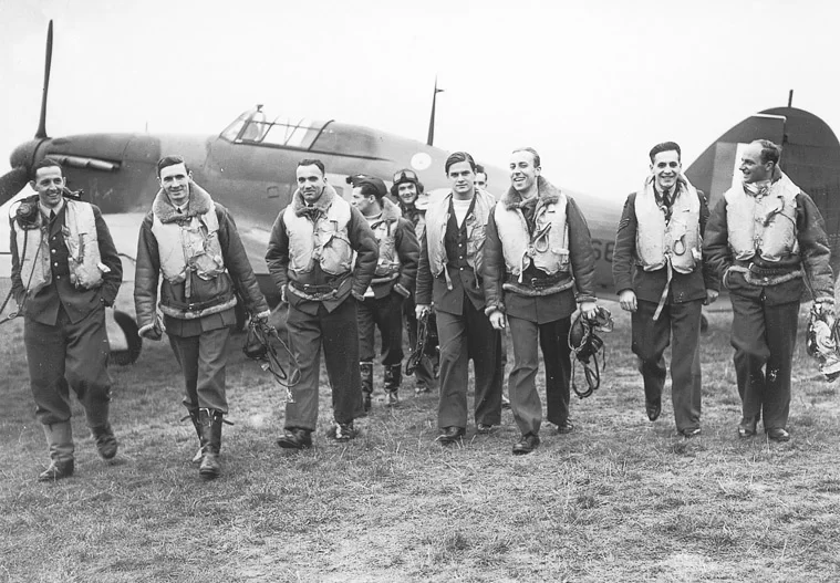 Zdjęcie przedstawia idących pilotów w mundurach, którzy trzymają zdjęte maski. Są uśmiechnięci. Za nimi stoi samolot bojowy.