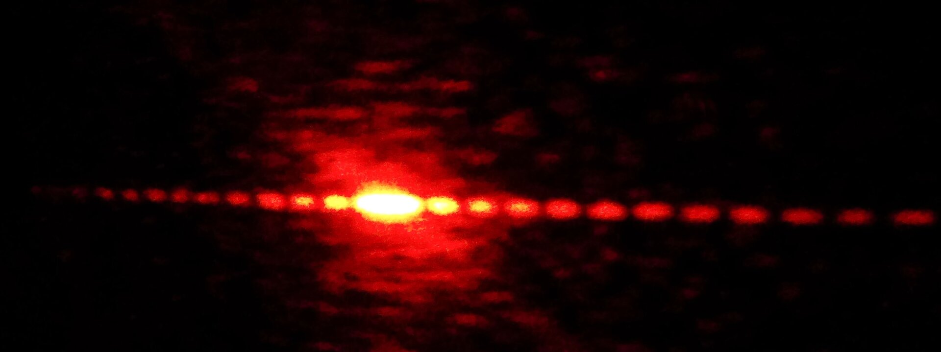 Rys. 1. Ilustracja przedstawia prążki powstałe przy przejściu światła monochromatycznego przez siatkę dyfrakcyjną. Światło monochromatyczne, to światło o jednej długości fali, a zatem i jednej barwie. Na czarnym tle pokazano czerwone punkty układające się na linii prostej biegnącej od lewego do prawego brzegu ilustracji. Środkowy punkt jest najjaśniejszy. Im dalej oddalone są punkty od punktu centralnego tym są one ciemniejsze, ponieważ natężenie światła w tych punktach jest coraz mniejsze. Prążki po lewej i prawej stronie od punktu centralnego rozmieszczone są symetrycznie.