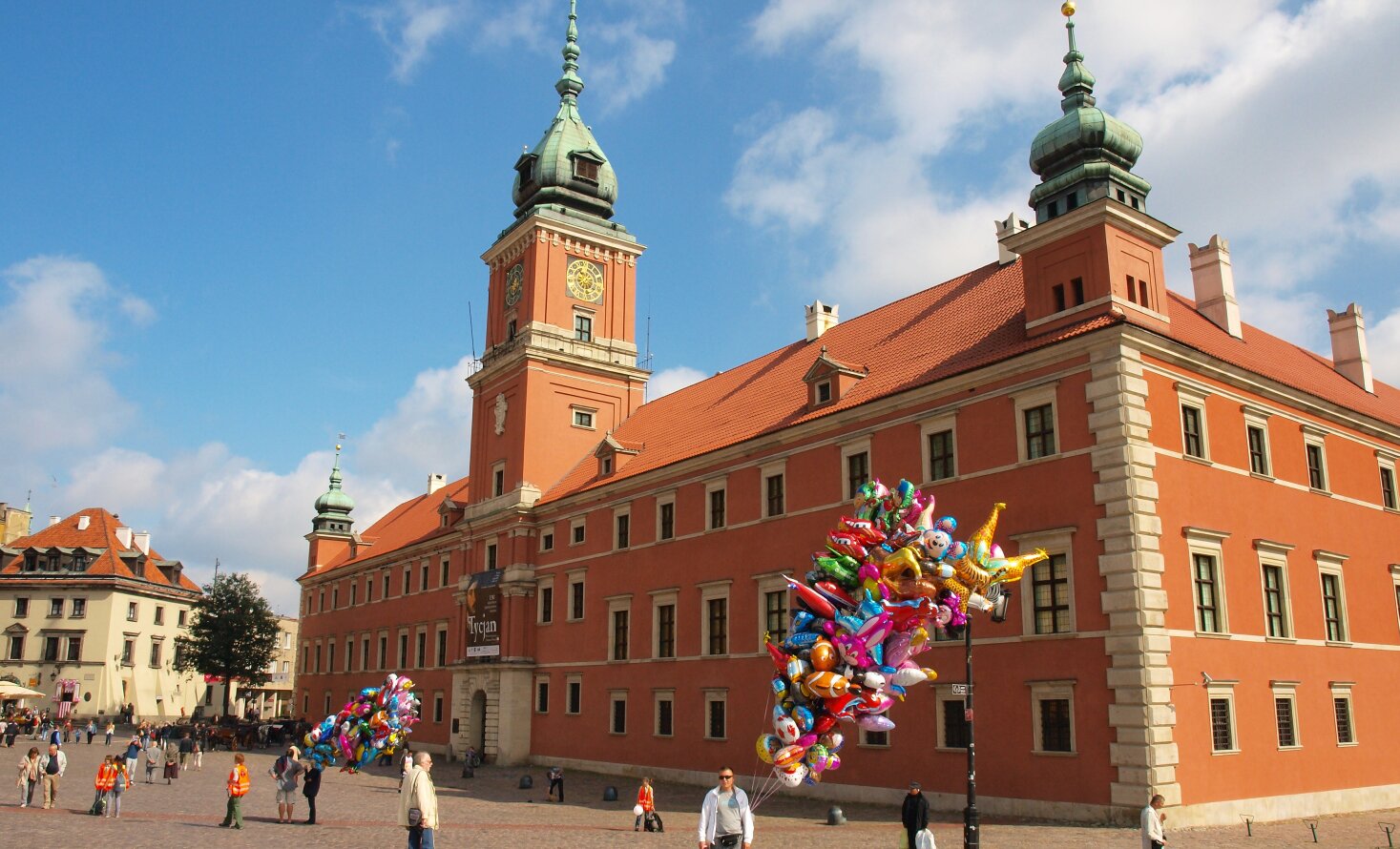 Fotografia barwna prezentująca fasadę warszawskiego Zamku Królewskiego  oraz fragment jego elewacji południowej. W centrum frontalnej, zachodniej części zamku znajduje się potężna wieża, zwana Wieżą Zygmuntowską. Ozdobiona okazałym zegarem z pozłacanymi wskazówkami. Pod nią znajduje się główne wejście do zamku. W narożach, po obu stronach, dwie mniejsze wieżyczki. Zamek pomalowany jest na świetlisty, pomarańczowy kolor. Ten okazały, monumentalny, trzypoziomowy  budynek mieści się na Placu Zamkowym.  Color photography presenting the facade of the Warsaw Royal Castle and a fragment of its southern façade. In the center of the front, west part of the castle there is a huge tower, called the Sigismund Tower. Decorated with a stately clock with gold-plated tips. Below it is the main entrance to the castle. In the corners, on both sides, two smaller turrets. The castle is painted in a bright orange color. This impressive, monumental, three-level building is located on Castle Square.