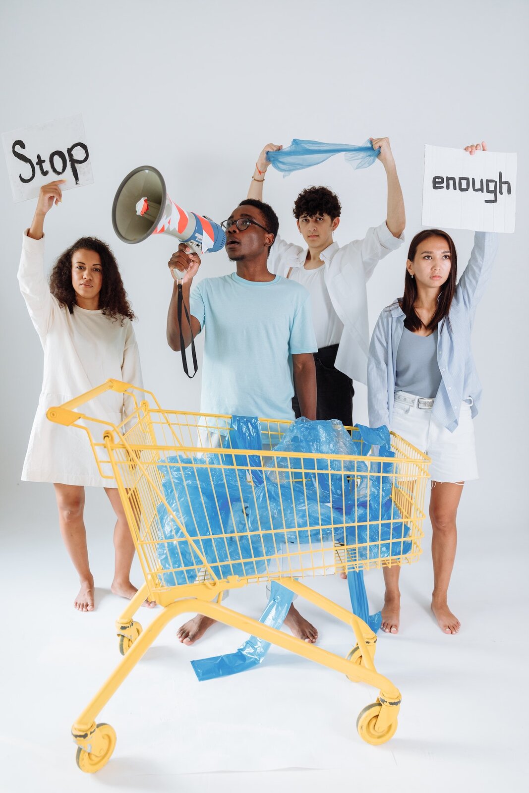 Zdjęcie przedstawia 4 osoby stojące obok siebie. Dwie kobiety stojące po bokach trzymają białe kartony z napisem  "STOP i "ENOUGH". Między nimi jest dwóch młodych mężczyzn, jeden stoi z megafonem przystawionym do otwartych ust, drugi trzyma nad głową foliowy worek, którymi jest wypełniony wózek zakupowy stojący z przodu przed całą grupą. Transkrypcja nagrań znajduje się poniżej.