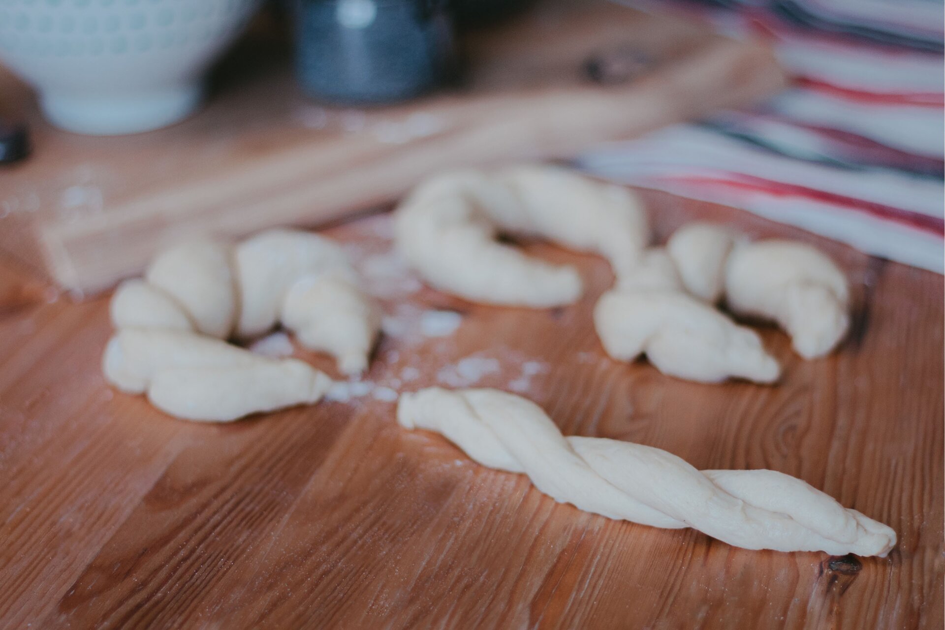 Zdjęcie przedstawia krok piąty formowania rogali węgierskich. Ujęcie wykonane jest od góry pod skosem. Na pierwszym planie w prawej dolnej części grafiki znajduje się spleciony wałek ciasta. Leży on na drewnianym ciemnobrązowym blacie. Nad nim w środkowej części grafiki dostrzec można niewyraźny zarys trzech uformowanych półokrągłych rogali. Dwa rogale znajdują się nad splecionym wałkiem, trzeci natomiast po jego prawej stronie. Na drugim planie w górnej części zdjęcia znajduje się niewyraźny zarys drewnianej deski na której stoi biała miseczka i szklany słoik. Pod deską leży biała ścierka w pasy o kolorze czerwonym i szarym.
