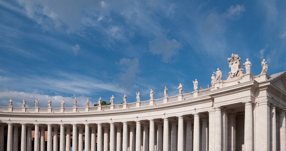 Półkolista kolumnada na placu św. Piotra w Watykanie, z długim rzędem wysokich kolumn i balustradą zwieńczoną licznymi marmurowymi posągami świętych. Nad centralną częścią widoczny jest ozdobny herb papieski. Nad architekturą rozciąga się jasne, błękitne niebo z lekkimi chmurami.