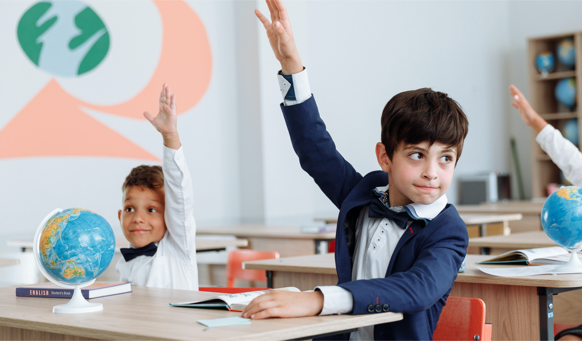 Zdjęcie przedstawia chłopców siedzących w ławkach szkolnych. Chłopcy podnoszą rękę, jak podczas zgłaszania się do odpowiedzi. Na ławkach stoją globusy.