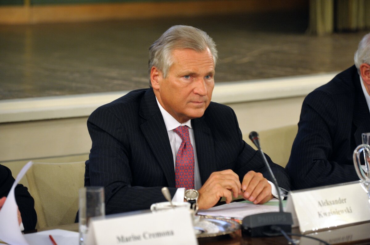 Kliknij żeby powiększyć Na zdjęciu znajduje się prezydent Aleksander Kwaśniewski. Mężczyzna z siwymi włosami siedzi za stołem. Za nim scena. Ma na sobie garnitur, a na prawej dłoni zegarek.