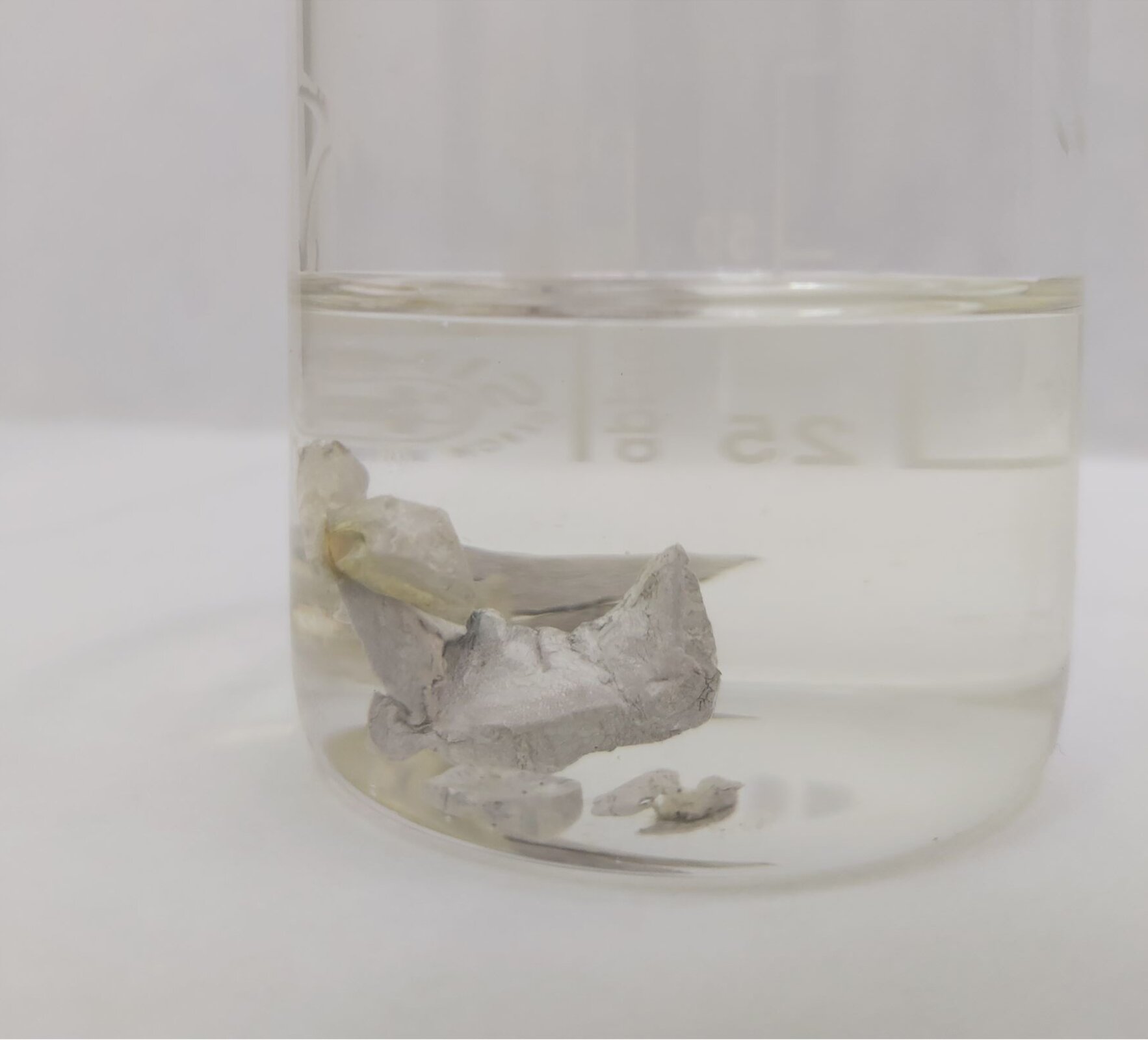 Na zdjęciu widoczny jest szklany pojemnik. W pojemniku znajduje się olej oraz zanurzone w nim kilka kawałków metalu o szarym kolorze.