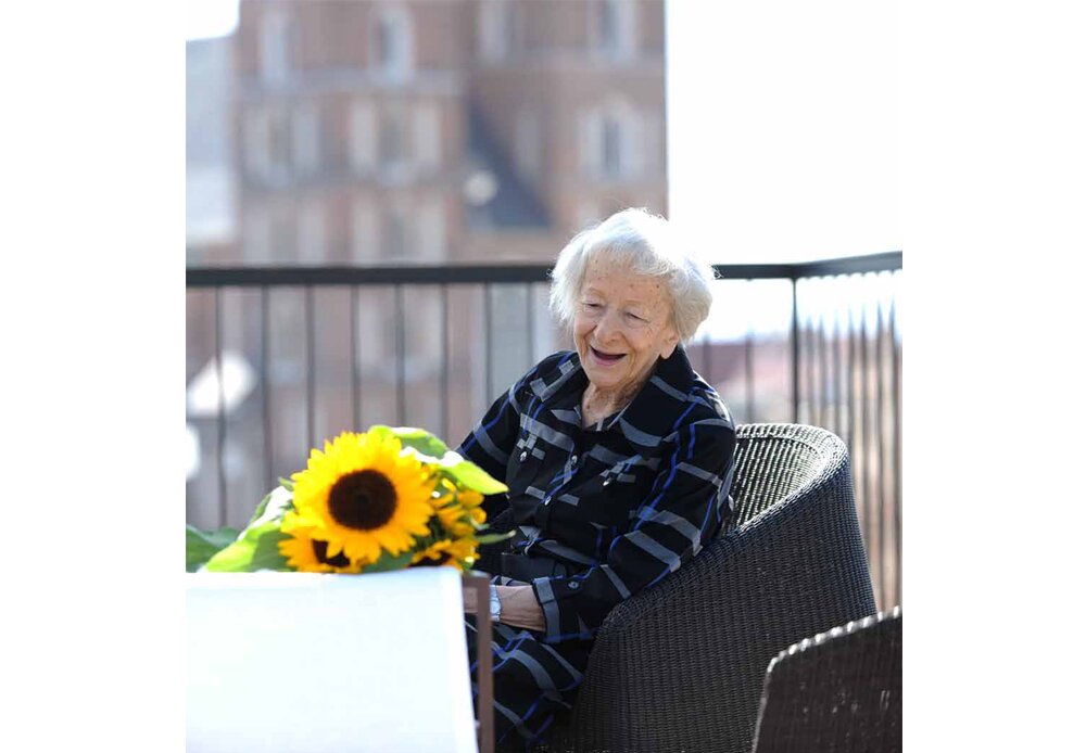 Fotografia barwna przedstawiająca Wisławę Szymborską. Siedzi na tarasie, obok niej leży bukiet słoneczników. Poetka uśmiecha się, jest słoneczny dzień.