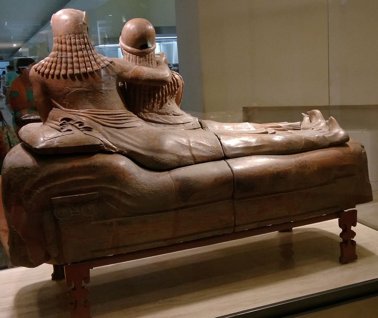 Zdjęcie przedstawia sarkofag wykonany z drewna. Jest on podłużny. Wieko przedstawia postacie dwóch wpółleżących osób – kobiety i mężczyzny. Są oni ukazani od strony swoich pleców. W górnej części znajdują się rzeźby ich tułowia, w dolnej części widać nogi. Wpółleżący mężczyzna obejmuje kobietę wpółleżącą przed nim.