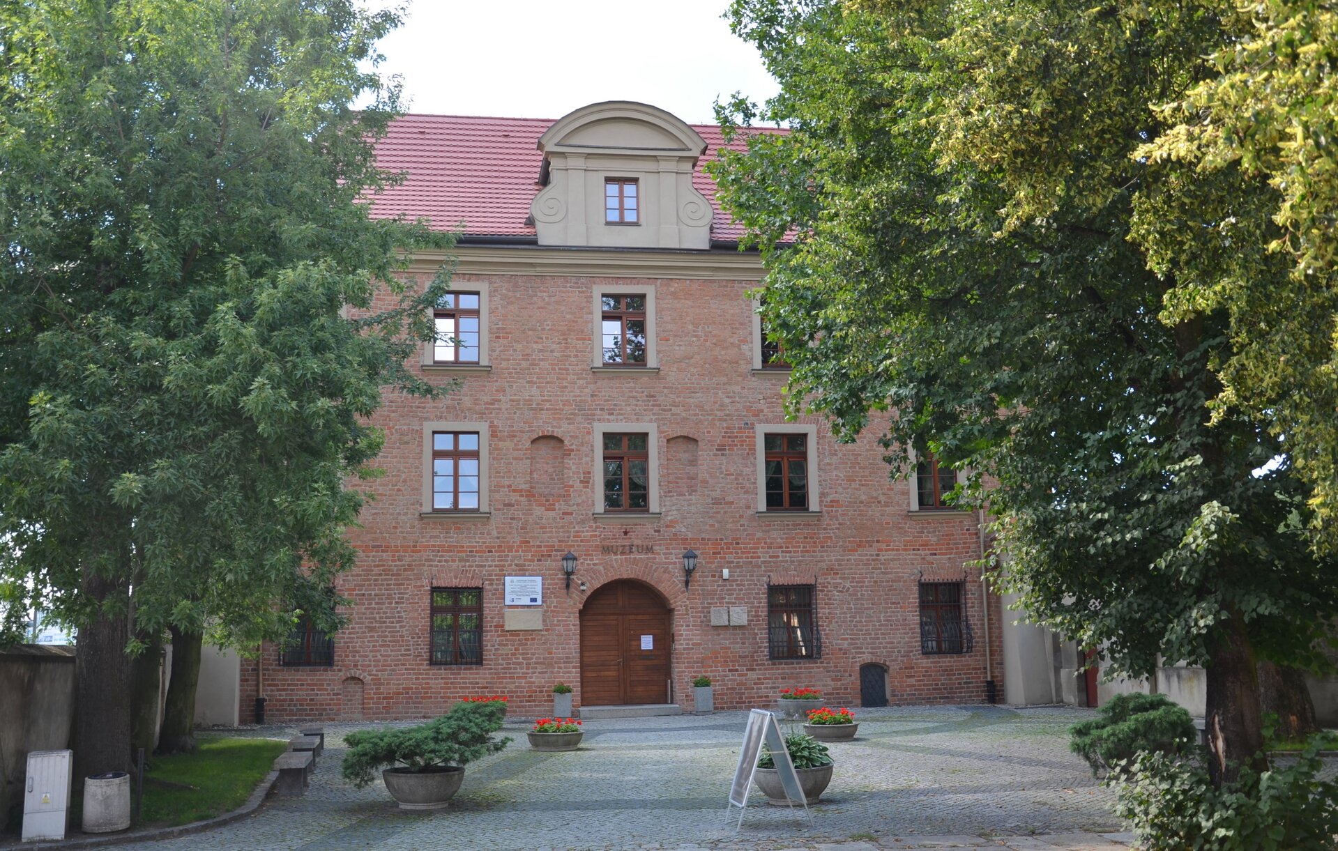 Ilustracja przedstawia akademię Lubrańskiego, która znajduje się na Ostrowie Tumskim w Poznaniu. Przed budynkiem widoczne są drzewa oraz kwiaty. Budunek zbudowano z czerwonej cegły. Okna są prostokątne z brązowymi ramami. Drzwi są półokrągłe, dachówka koloru czerwonego.