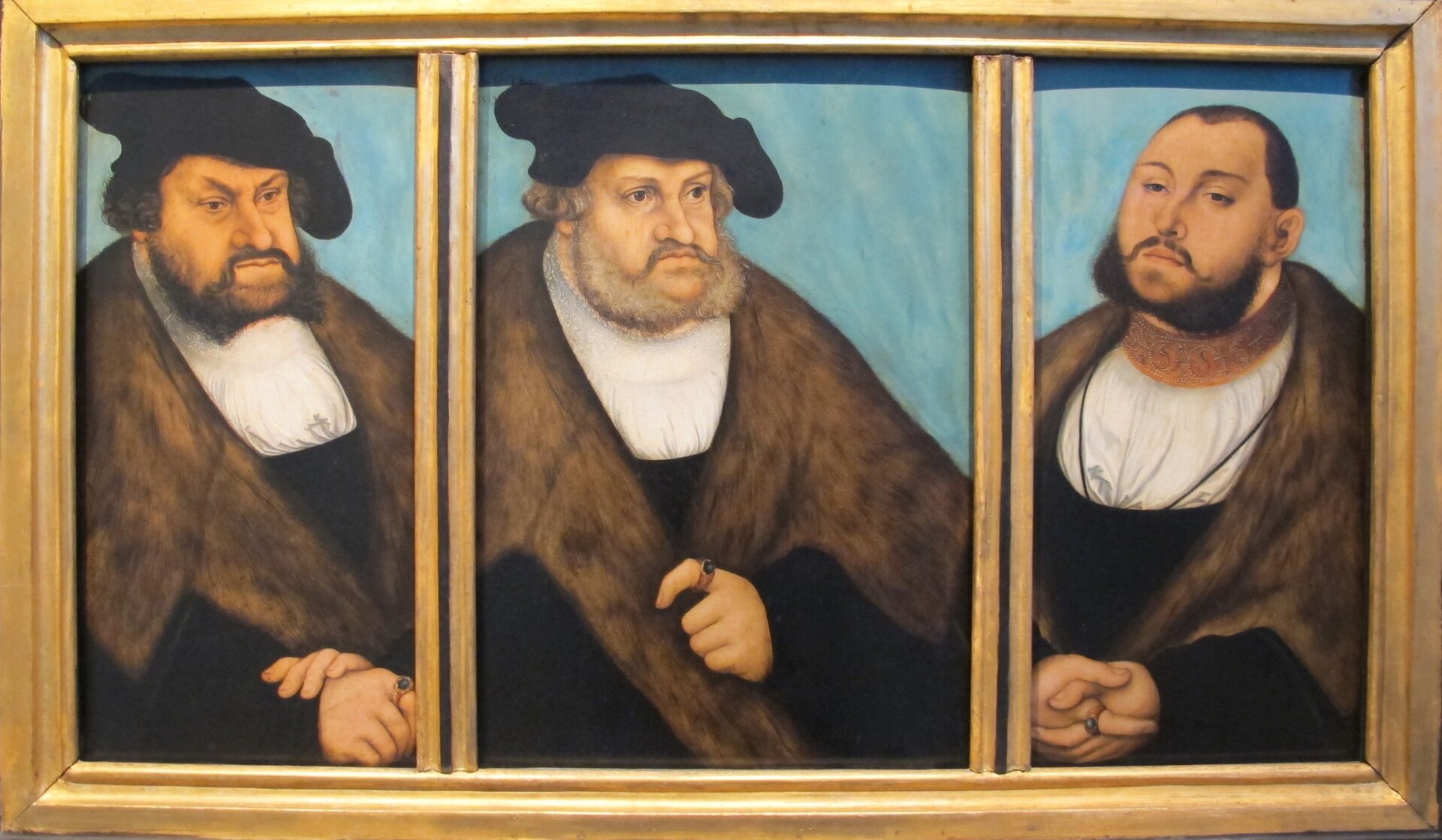 Obraz przedstawia portrety trzech mężczyzn, które zostały ustawione jeden obok drugiego. Mężczyźni są w średnim wieku i ubrani w ten sam sposób: biała koszula oraz czarny płaszcz z futrem przy szyi. Dwóch mężczyzn, znajdujących się z lewej strony, ma na głowach czarne czapki. Mężczyźni mają dłuższy zarost i wąsy.