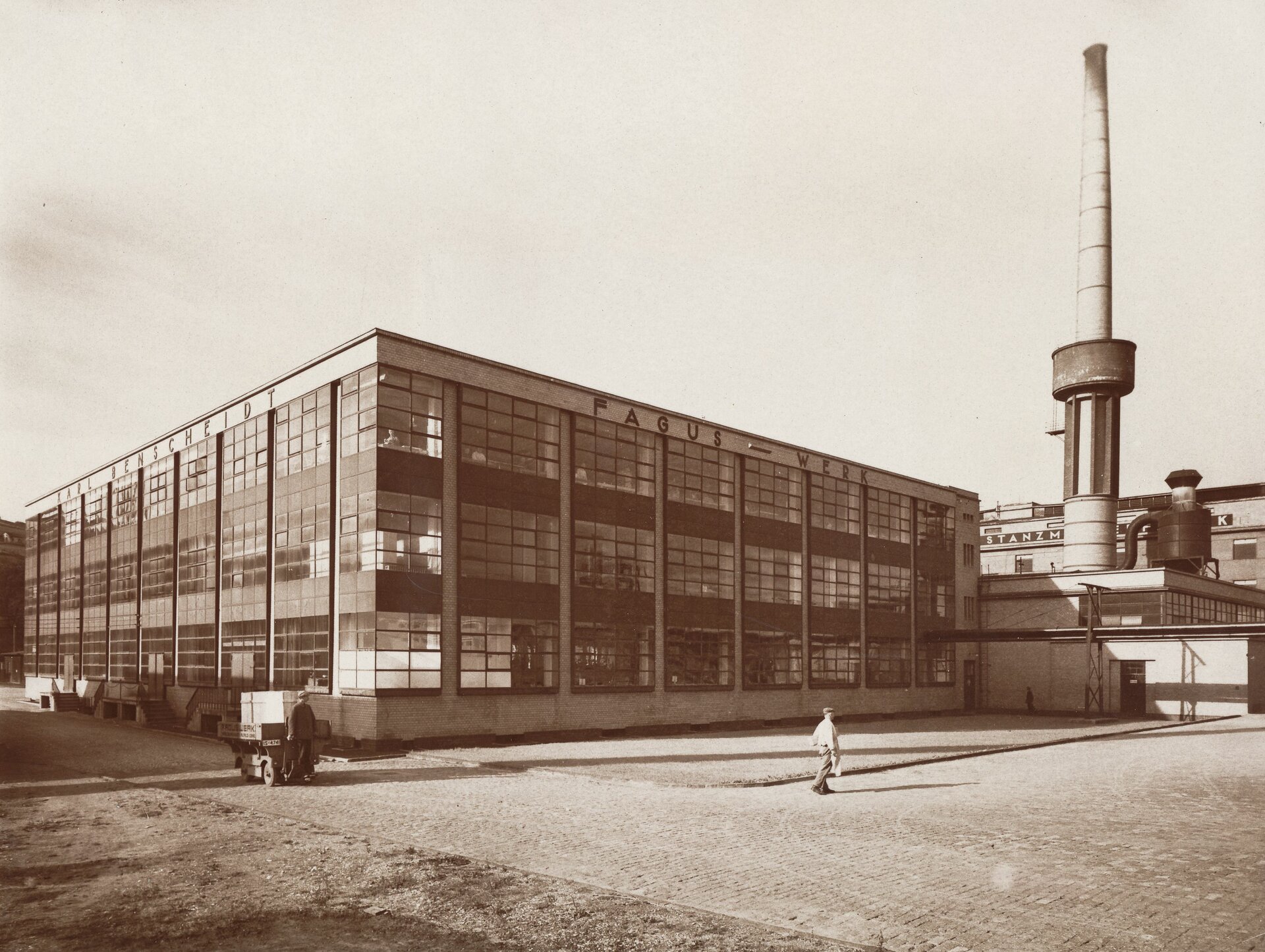 Ilustracja przedstawia czarno białą fotografię Waltera Gropius i Adolfa Meyer „Fabryka Fagus”. Na pierwszym planie widać dwie idące osoby, w tle znajduje się budynek fabryki z przeszklonymi ścianami. Po prawej stronie znajduje się wysoki komin i inne budynki.