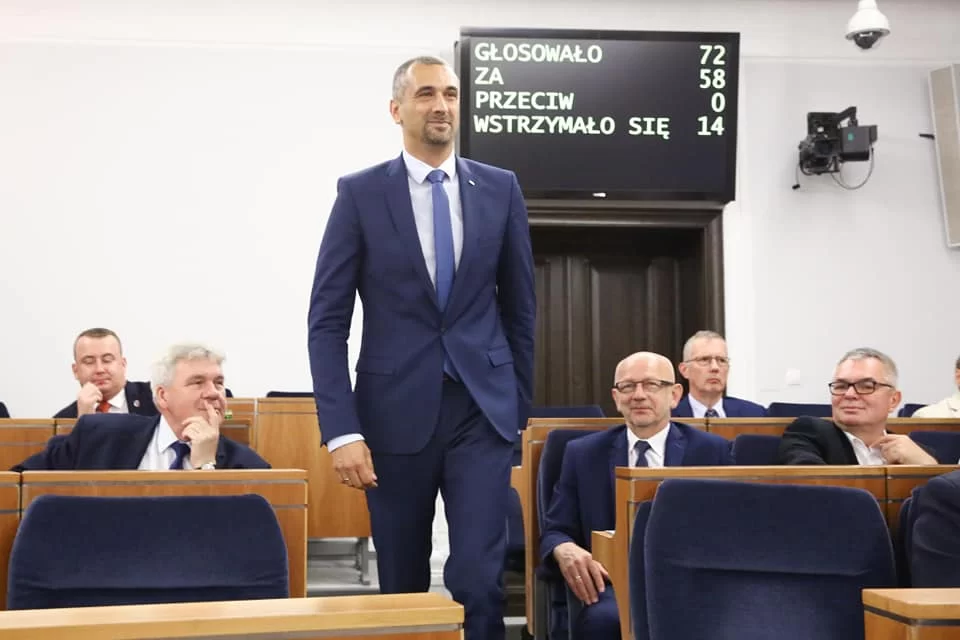 Ilustracja przedstawia senatora idącego środkiem sali. Jest to dość młody wysoki, szpakowaty mężczyzna. Ubrany w elegancki garnitur, koszulę oraz krawat. Po obu stronach senatora siedzą inni mężczyźni w drewnianych ławkach.