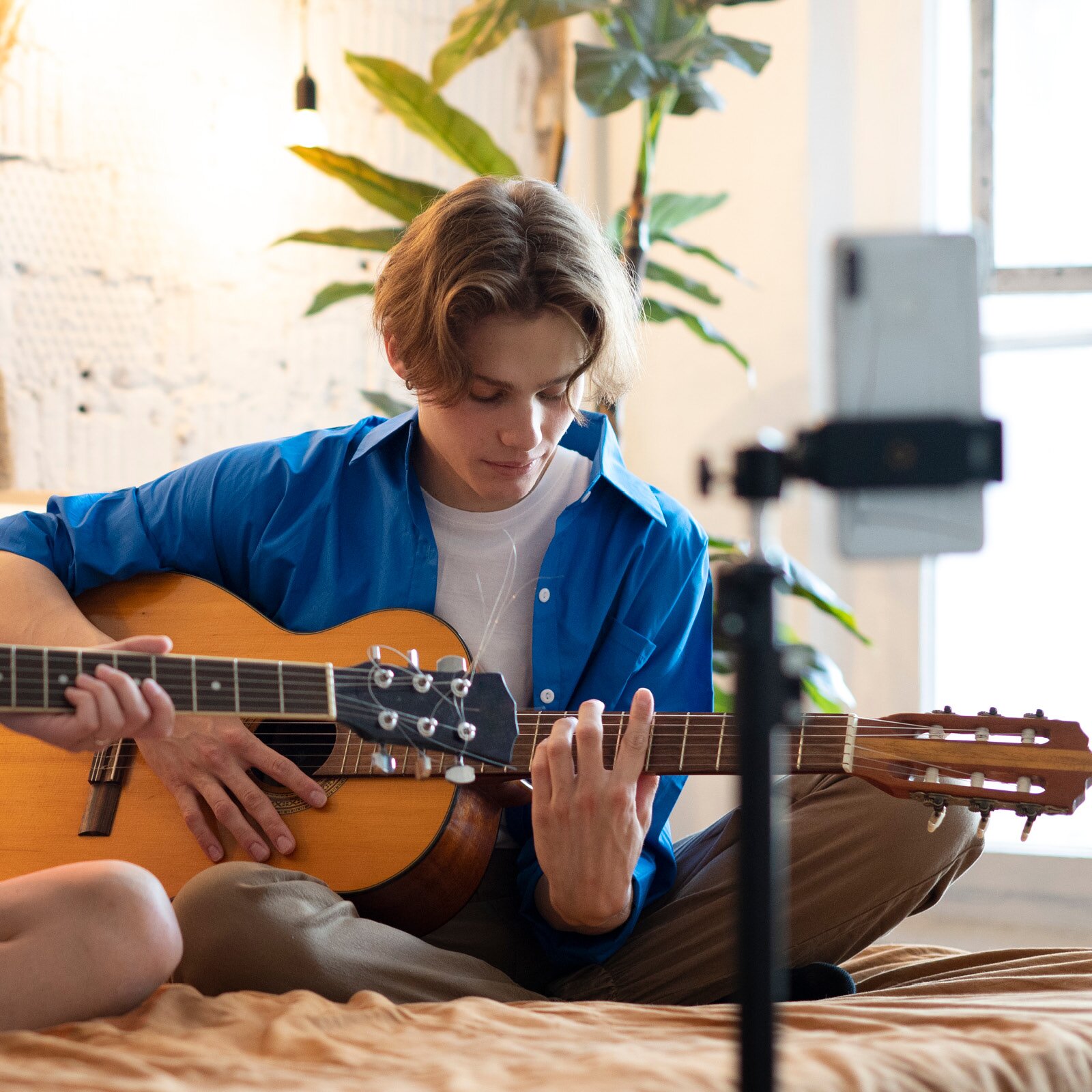 Zdjęcie przedstawia nastoletniego chłopaka siedzącego na łóżku i grającego na gitarze klasycznej.