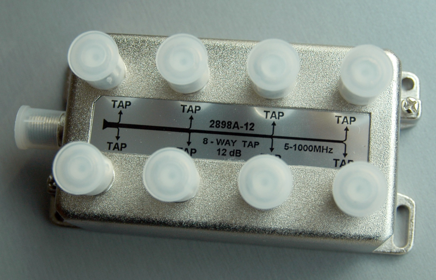 Ilustracja przedstawia odgałęźnik 8 tap 2. To urządzenie stosowane w telewizji kablowej. Ma kształt płaskiego pudełka, wykonany jest z metalu. Na wierzchu znajdują się napisy informujące o jego przeznaczeniu. Na górze odgałęźnika widocznych jest także osiem otworów, z których każdy posiada ochronne nakładki z tworzywa. Odgałęźnik umożliwia podłączenie większej liczby odbiorników.