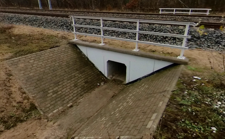 Zdjęcie przedstawia przepust. Jest to niski tunel biegnący pod torem kolejowym. Przed wejściem do tunelu ściany rowu do niego prowadzącego są wymurowane i pochylone, tworząc w przekroju kąt rozwarty. Nad wejściem do przepustu widoczna jest barierka.