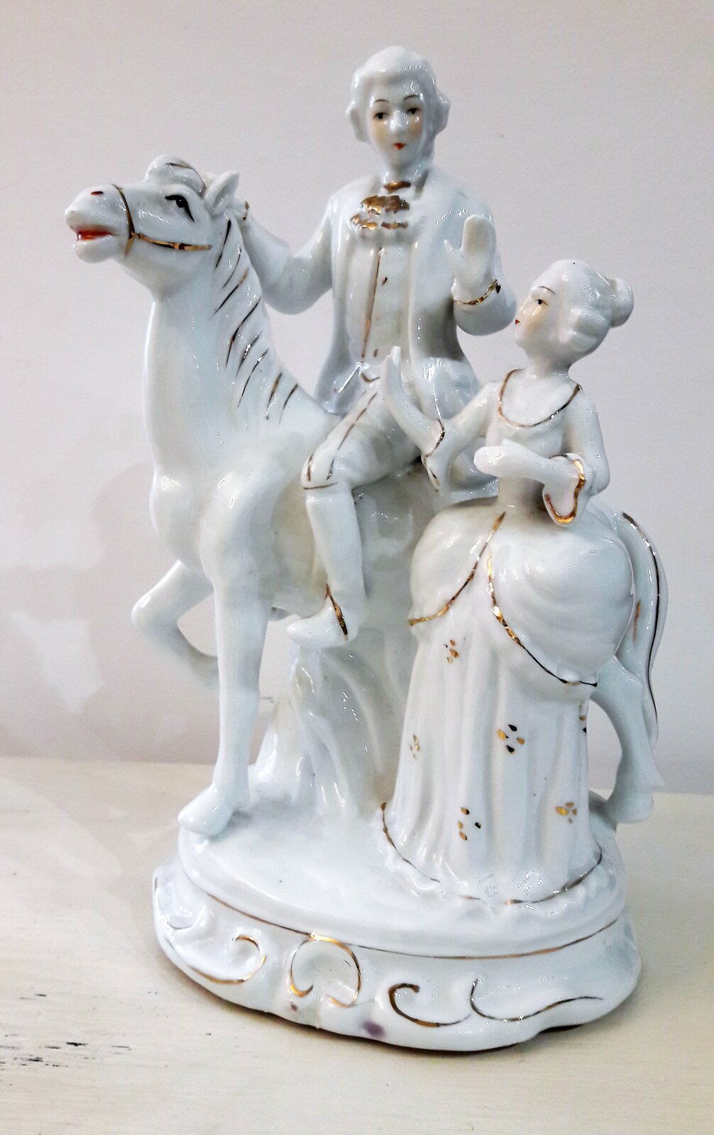 Ilustracja przedstawia porcelanową figurkę manufaktury bawarskiej, przedstawiającą mężczyznę na koniu i stojącą obok kobietę. Postacie mają na sobie bogate stroje. Kobieta zwrócona jest w stronę mężczyzny, prawdopodobnie rozmawiają. Mężczyzna jedną ręką trzyma konia, drugą gestykuluje. Łeb konia uniesiony jest w górę.