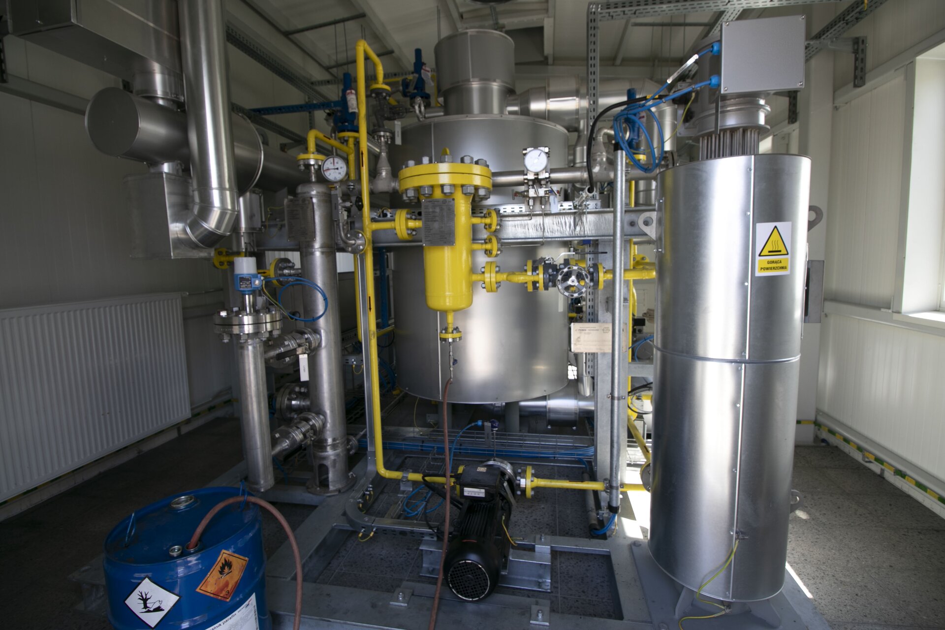 Na ekranie widoczny jest kondensator. Kondensator wykorzystuje chłodzenie wodne lub powietrze w celu skroplenia gorących par w ciecz.W centralnej części zdjęcia znajduje się duży cylinder. Z jego góry poprowadzone są rury. W połowie jego wysokości w orientacji poziomej poprowadzona jest belka. Z prawej strony zdjęcia znajduje się cylindryczny zbiornik o mniejszym promieniu niż ten na środku. W jego górnej części widoczne są pionowe druty, a za pomocą niebieskich przewodów połączony jest z resztą układu. W lewej części zdjęcia znajdują się dwie pionowe rury oraz jedna pozioma umieszczona nad nimi. Pierwsza pionowa rura dołączona jest do poziomej. Przed dużym cylindrem i belką znajduje się mały żółty cylinder, z którego odchodzi wiele rur. Rury małego cylindra połączone są z drugą pionową rurą obok, której znajduje się ciśnieniomierz, z czarną pompą znajdującą się na ziemi, zaworem oraz innymi elementami układu. Na pierwszym planie zdjęcia znajduje się niebieska beczka z symbolem łatwopalnej substancji. Jest ona połączona wężem z żółtym cylindrem.