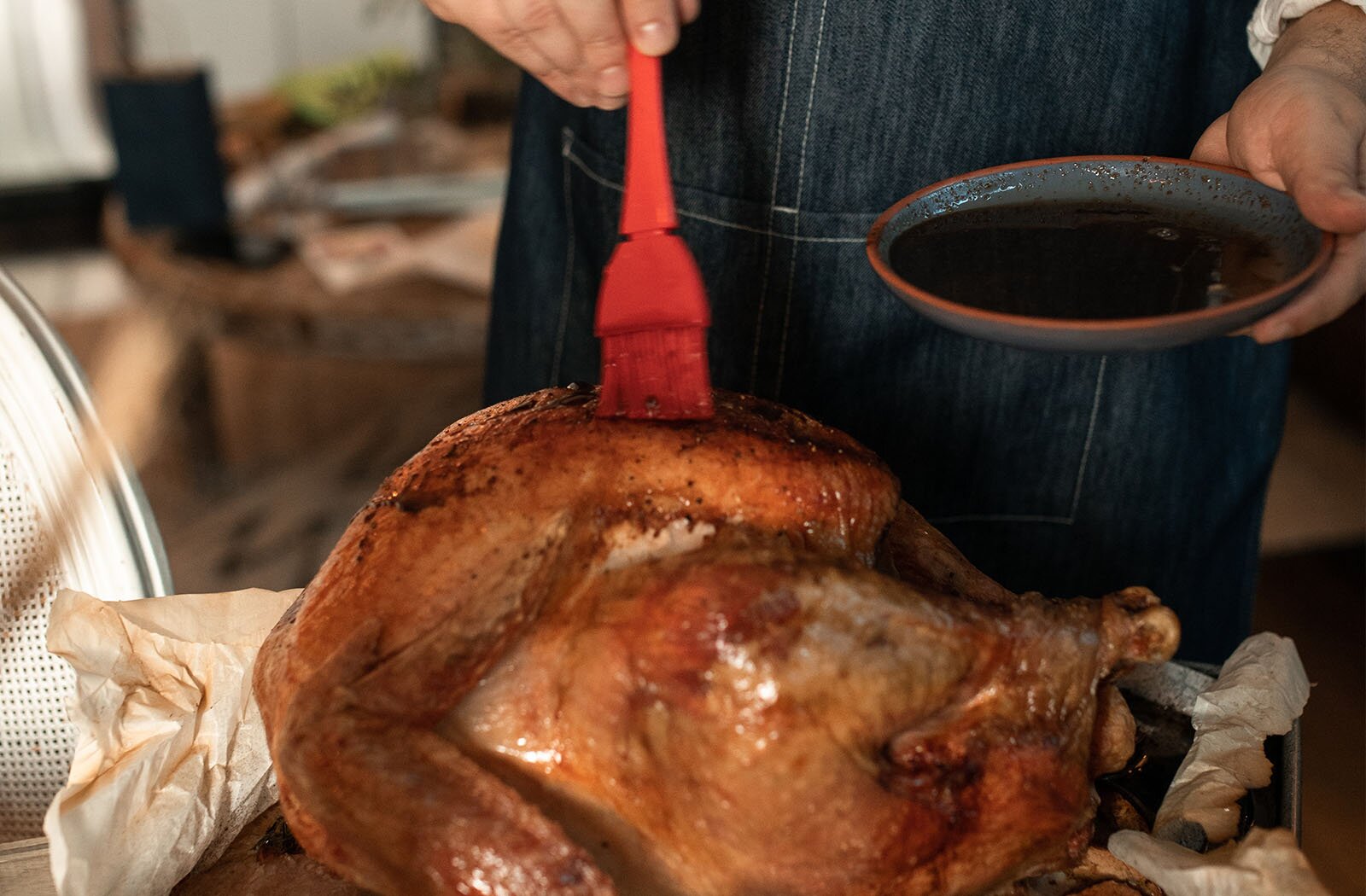 Zdjęcie przedstawia leżącego na pergaminie upieczonego indyka, ze spieczoną skórką, która jest posypana ziołami. W głębi widoczna jest postać, która w prawej ręce trzyma czerwony pędzel kuchenny, zaś w lewej miseczkę z ciemnym płynem. Postać - za pomocą pędzelka – rozprowadza sos na indyku. 