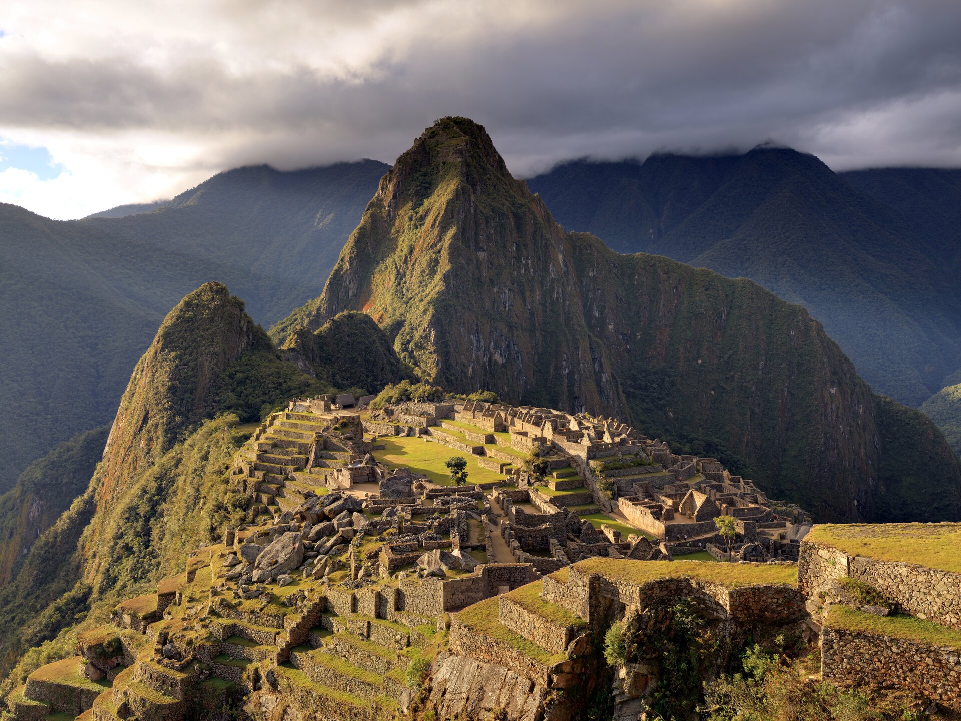 Fotografia barwna przedstawiająca Machu Picchu. Na wzniesieniu liczne zabudowania. Wokół góry. 
