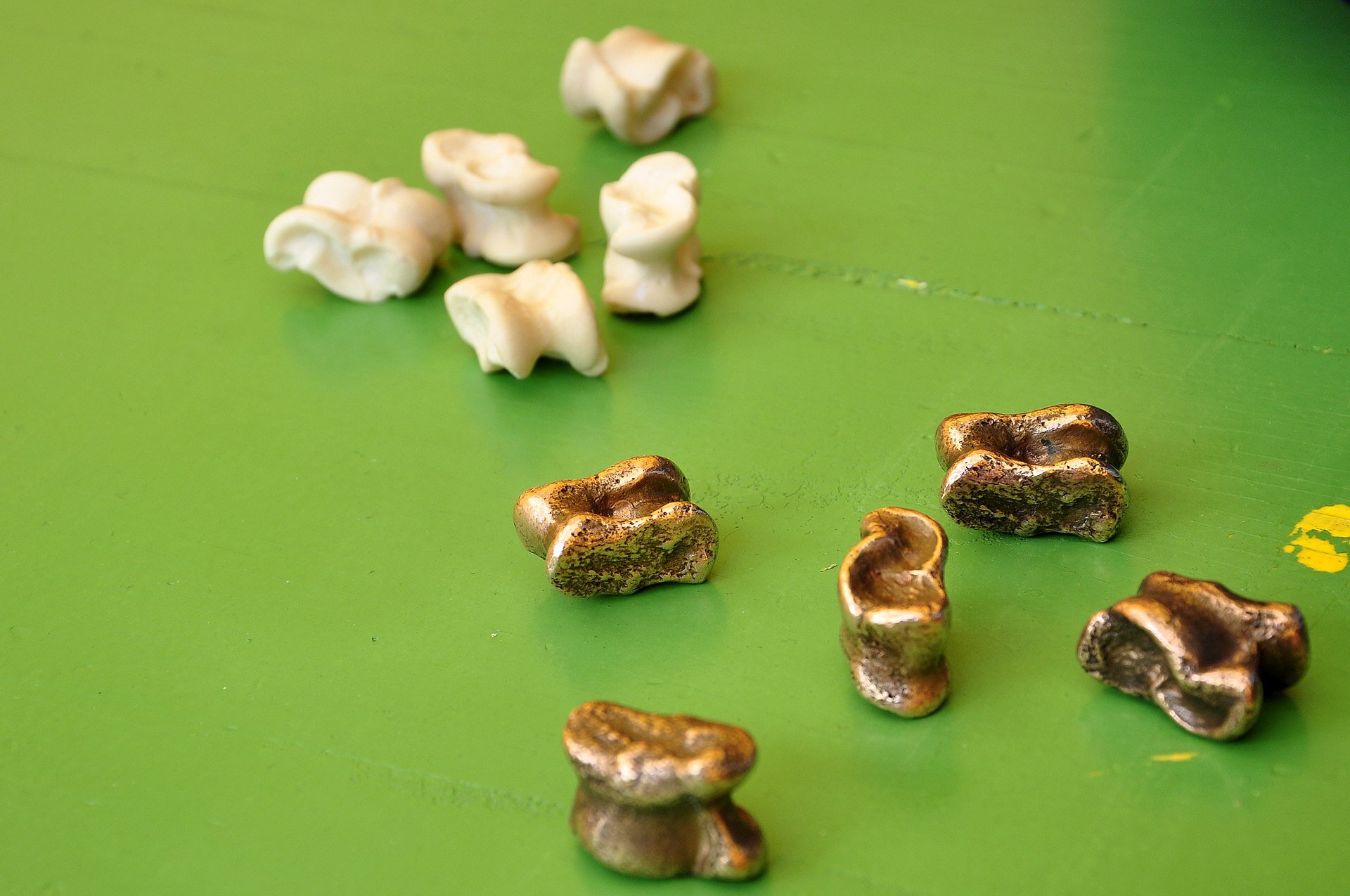 Zdjęcie przedstawia ułożone na jasnozielonym stole dziesięć niewielkich elementów, w kolorach białym oraz złotym. Ich kształty są nieregularne, jednak przypominają pofalowane owale ze żłobieniami.