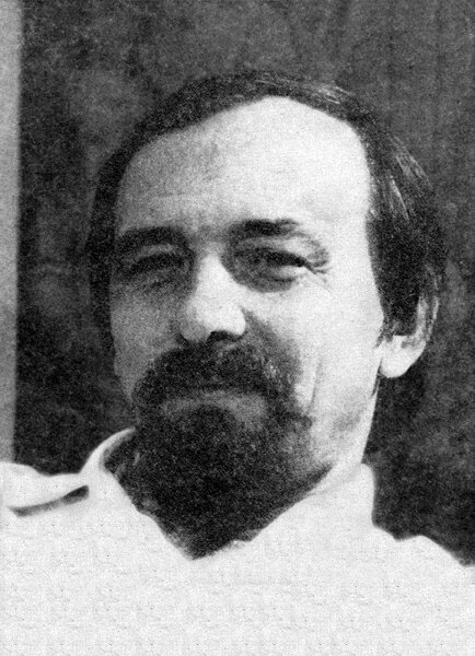 Zdjęcie przedstawia portret mężczyzny w sile wieku. Postać ukazuje lewy półprofil. Mężczyzna jest zaprezentowany od piersi w górę. Twarz mężczyzny jest owalna. Ma on wysokie czoło i nieznaczne zakola. Włosy są ciemne, proste, zaczesane na prawą stronę. Brwi mężczyzny są krzaczaste, ciemne. Postać ma oczy zwężone w uśmiechu, otoczone zmarszczkami mimicznymi. Mężczyzna patrzy na nas. Ma jastrzębi nos. Jego twarz zdobią gęste wąsy i broda na podbródku. Policzki są gładko ogolone. Mężczyzna jest ubrany w jasną koszulę.