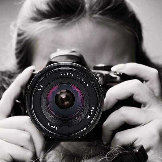 Zdjęcie przedstawia fotografującą dziewczynę. Aparat z otwartą przysłoną zajmuje centralne miejsce, jest trzymany oburącz przez dziewczynę, która patrzy przez obiektyw. Za aparatem widoczne jest tylko jej czoło i włosy. 