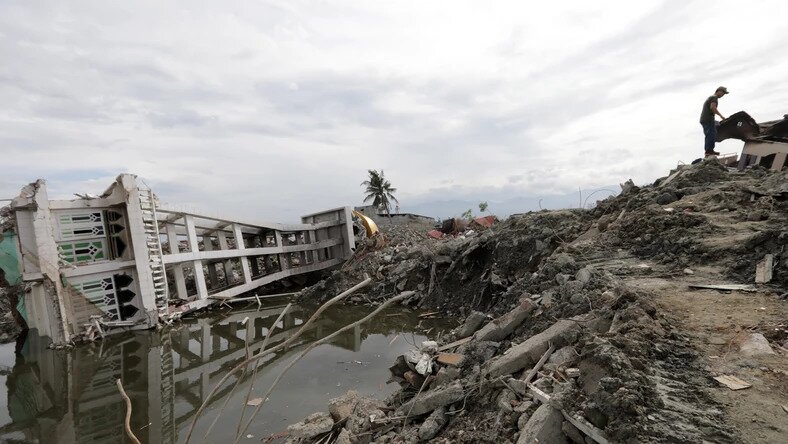 Zdjęcie przedstawia Indonezję po tsunami. Z prawej strony znajduje się wydeptana ścieżka. Z lewej strony tworzy niewielką skarpę, za którą jest zebrana woda. Na wodzie leży przewrócony wysoki budynek. 