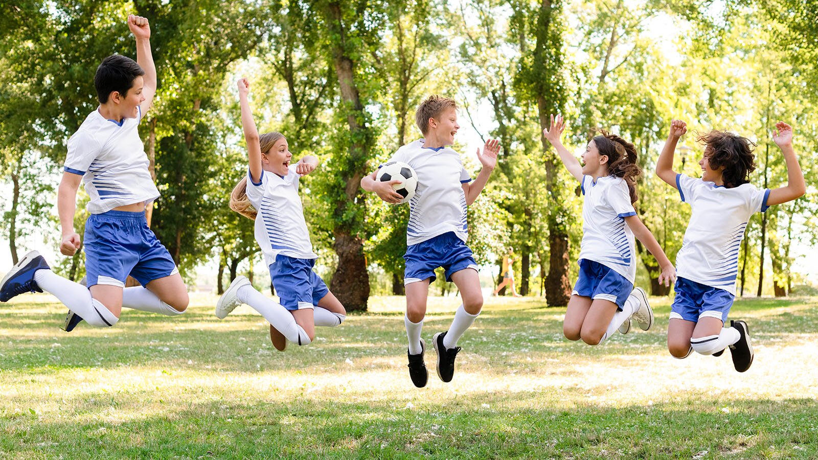 Zdjęcie przedstawia dzieci w wieku szkolnym skaczące na trawie w parku. Wszyscy ubrani są w stroje sportowe, krótkie spodenki i koszulki na krótki rękaw. Chłopiec w środku grupy trzyma w dłoniach piłkę. 