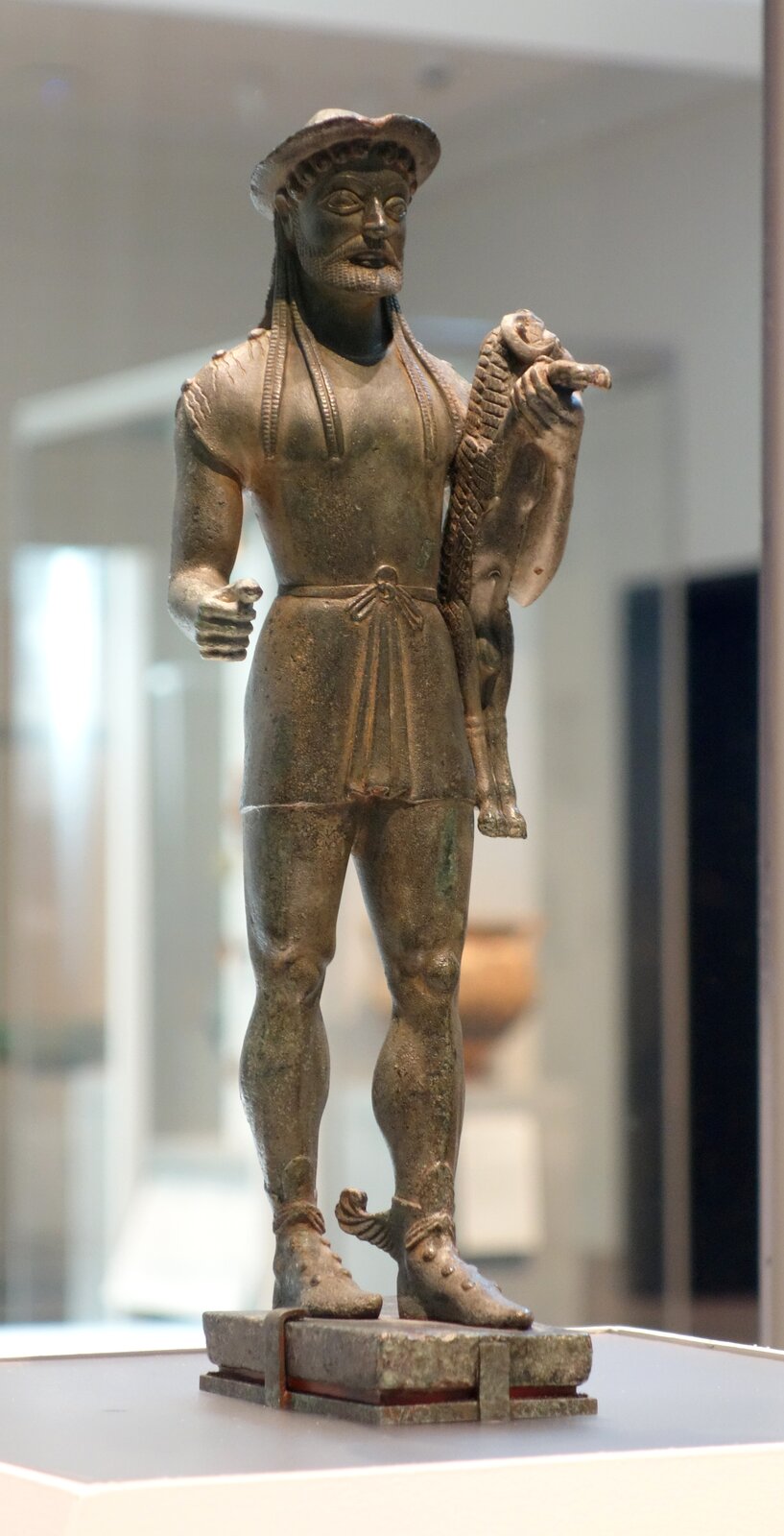 Brązowa rzeźba ukazuje młodego mężczyznę identyfikowanego jako Hermes, niosącego barana pod lewym ramieniem. Postać stoi frontalnie, z wyważoną, spokojną postawą i subtelnym archaicznym uśmiechem. Anatomia ciała jest uproszczona, lecz czytelna, z zaznaczonymi mięśniami i symetrią typową dla późnego okresu archaicznego. Powierzchnia brązu jest gładka, z delikatną patyną i nielicznymi ciemniejszymi plamami, a całość ma charakter rytualny i ofiarny.