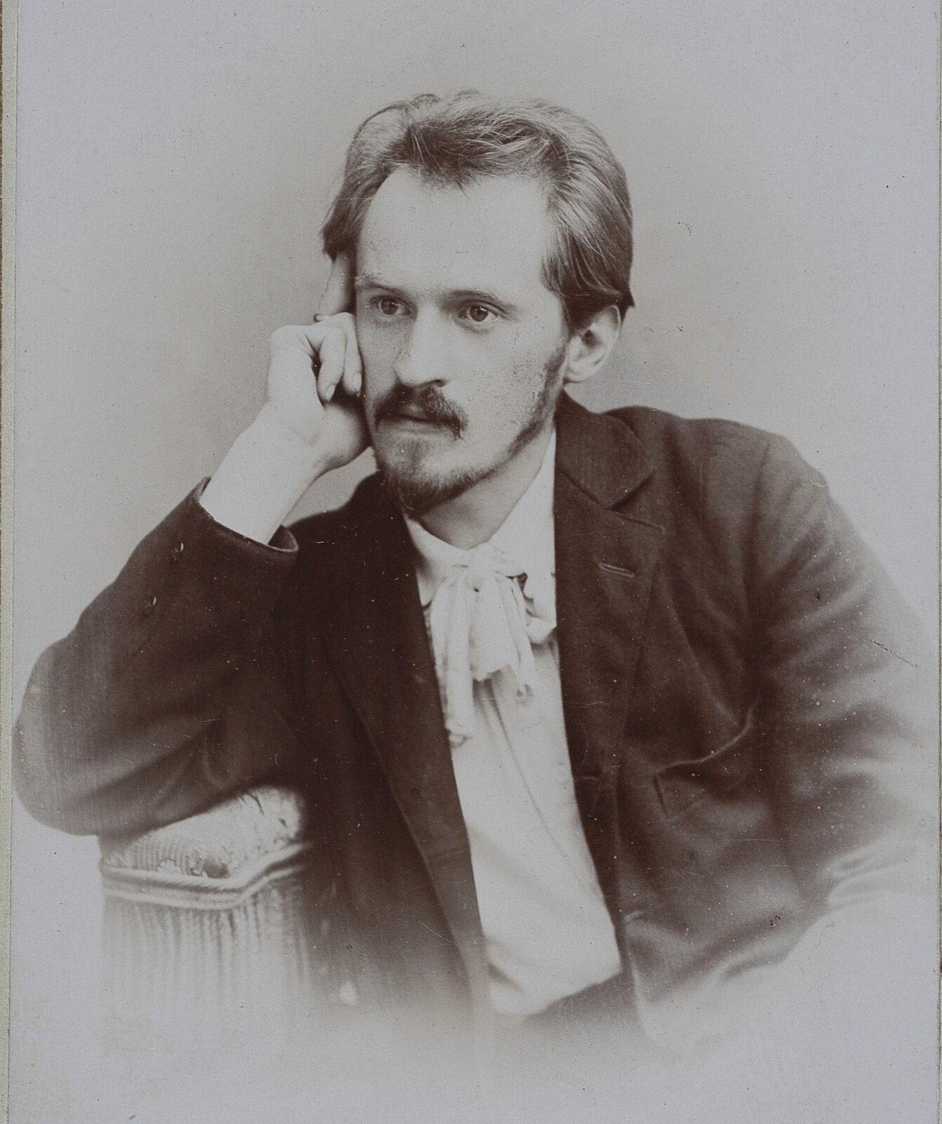 Zdjęcie przedstawia portret młodego mężczyzny. Jego postać widoczna jest od pasa w górę. Mężczyzn siedzi wsparty prawą ręką na fotelu. Prawą dłonią wspiera głowę. Mężczyzna ma pociągłą twarz. Ma krótką brodę i wąsy. Jego włosy są proste, zaczesane ku górze. Mężczyzna ubrany jest w koszulę i marynarkę.