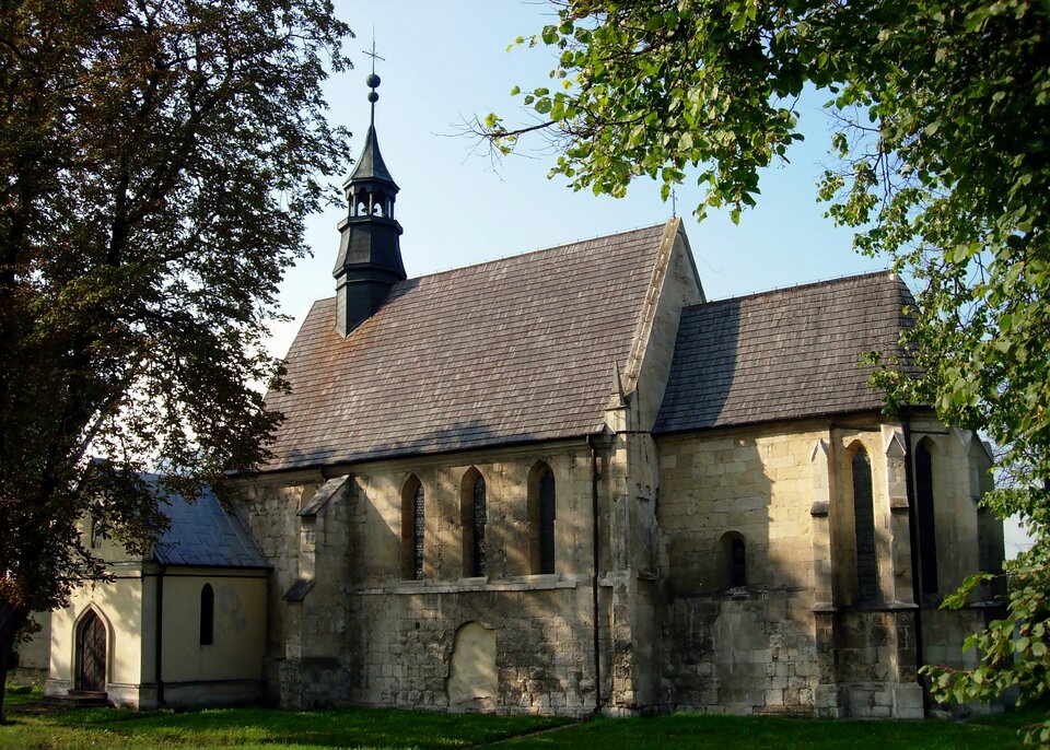 Fotografia przedstawia budynek zbudowany z kilku brył. Ma wysokie okna, gładkie ściany i spadzisty dach, na którym widać wieżę z dzwonem w środku, zakończoną krzyżem. 