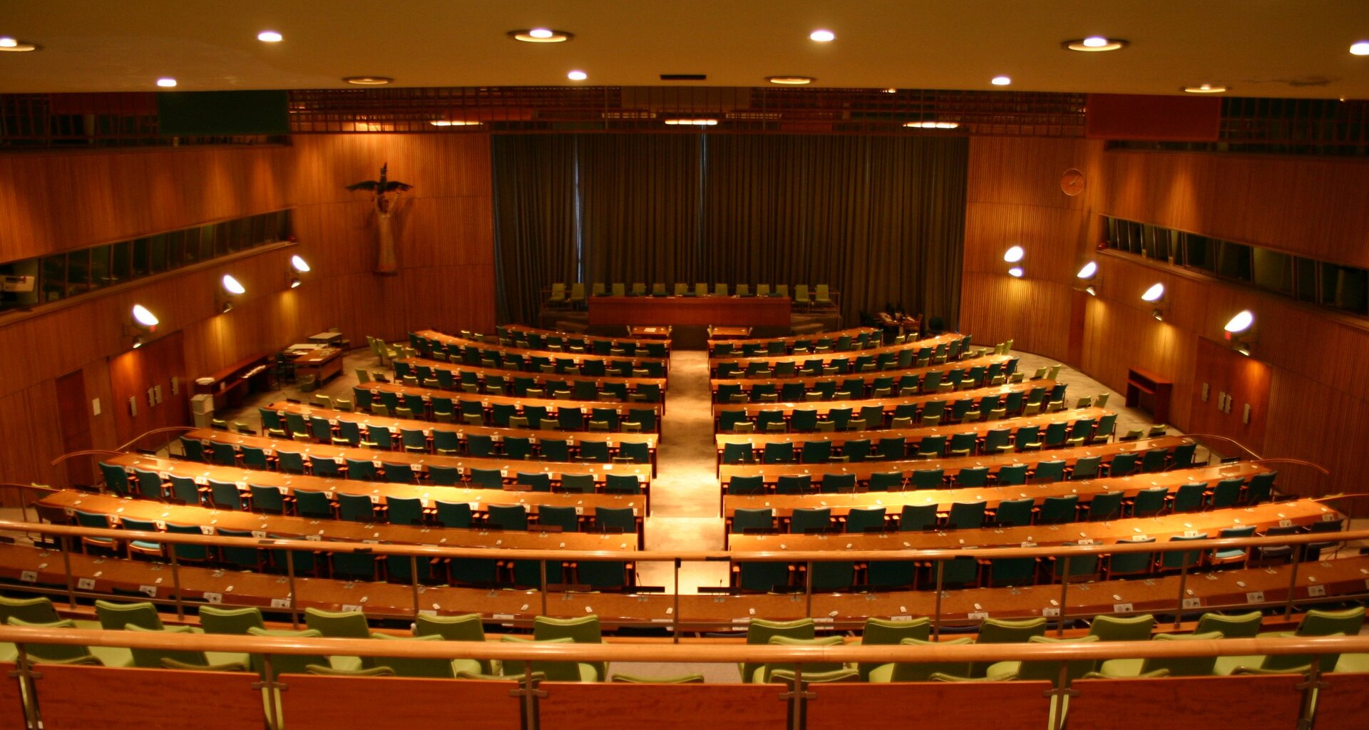 Zdjęcie przedstawia dużą aulę. W tle widoczne jest podium, na którym stoi długi stół, a za nim ustawione są fotele. W tle widoczne są długie brązowe zasłony. Na pierwszym planie widoczne są kaskadowo ustawione fotele oraz drewniane blaty. Na ścianach bocznych zamocowano halogeny. Oświetlenie punktowe widoczne jest na suficie.