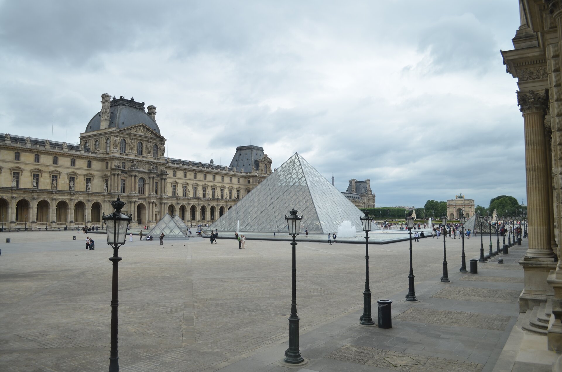 Zdjęcie wykonane w Paryżu w pochmurny dzień przedstawia przezroczystą budowlę w kształcie piramidy. Po lewej i prawej stronie piramidy stoją beżowe budynki z wieloma oknami z szarym dachem. W oddali za piramidą stoi Łuk Triumfalny.