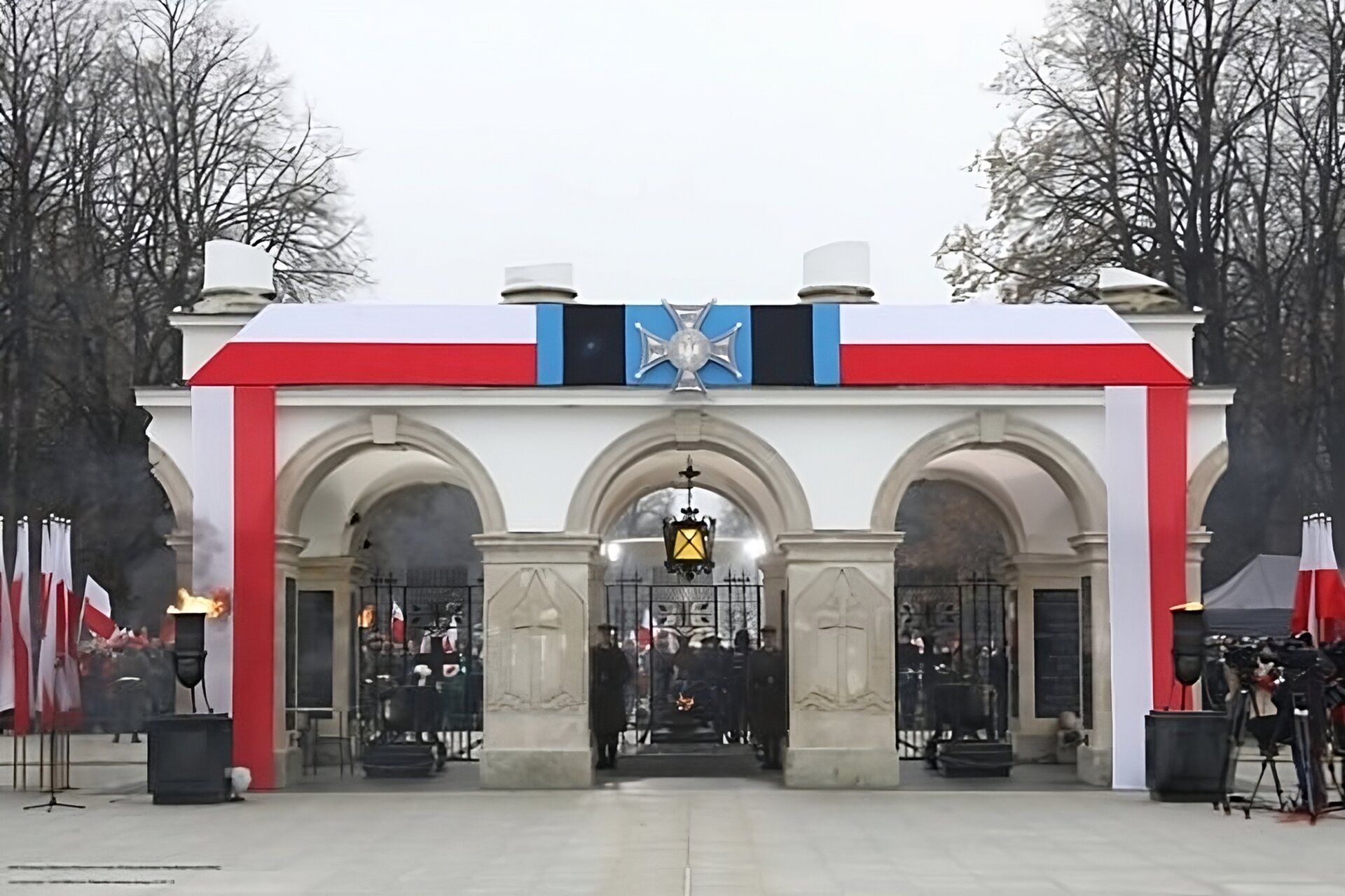 Zdjęcie przedstawia udekorowany biało‑czerwonymi pasami materiału Grób Nieznanego Żołnierza w Warszawie 11 listopada 2018 roku dla uczczenia 100‑lecia odzyskania przez Polskę niepodległości. Konstrukcja składa się z trzech arkad. Centralnie, nad główną arkadą grobu, zawieszono imitację Krzyża Walecznych. Po obu stronach grobu ustawiono stojaki z flagami narodowymi oraz płonące znicze. W centrum widać świecącą, podwieszaną pod arkadami latarnię. Poniżej dwóch żołnierzy - wartowników, stojących na baczność. W tle, za arkadami, widoczne tłumy ludzi z flagami państwowymi. Na obu krawędziach fotografii widoczne są bezlistne drzewa, niebo zamglone. W dolnej części fotografii płyty chodnikowe.