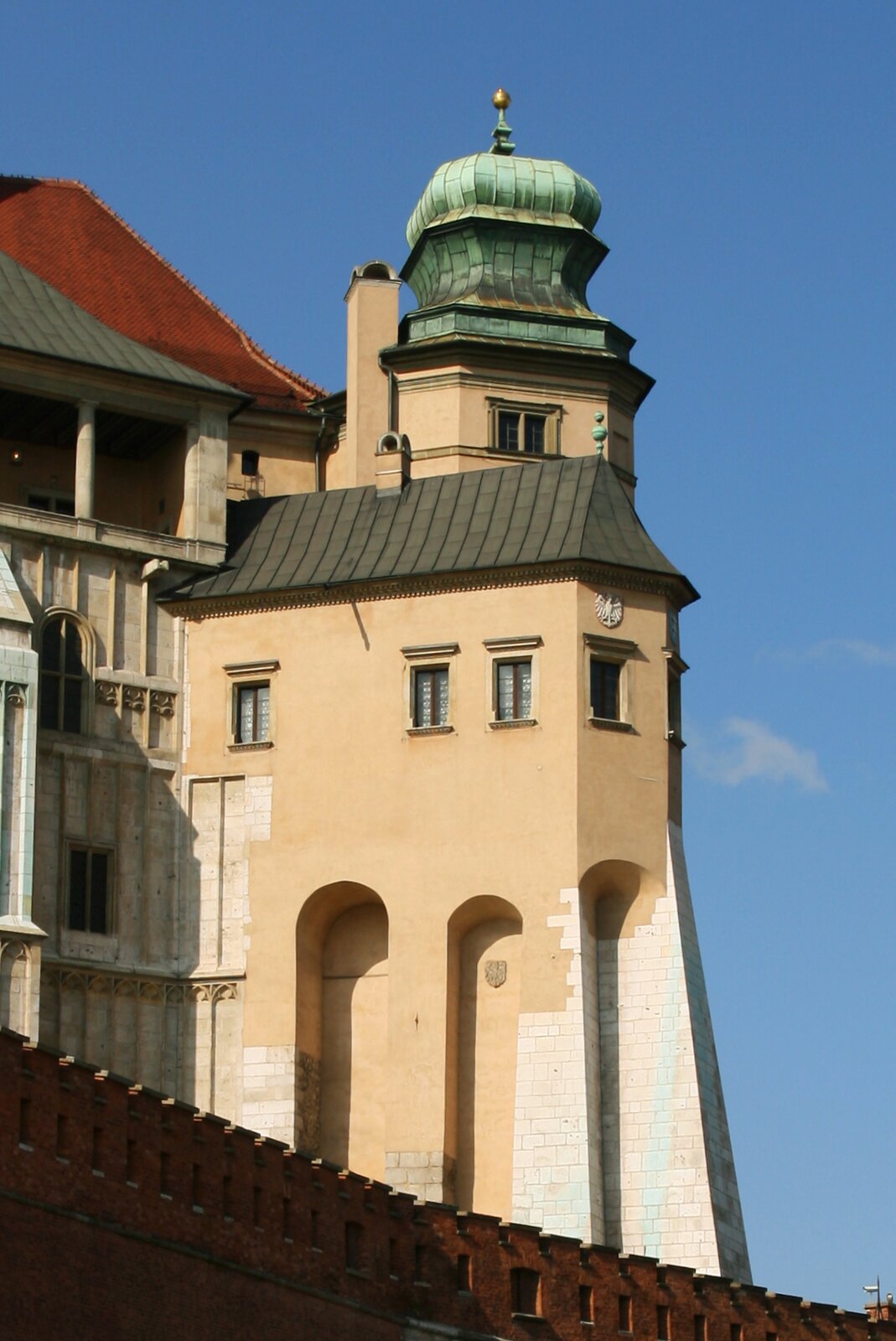 Na zdjęciu wieża Kurza Stopka na zamku wawelskim. Widać trzy łukowe wnęki, ciągnące się do połowy wieży. Nad wnękami pięć prostokątnych  okien. Na drugim od prawej herb z orłem. nad oknami spadzisty dach a nad nim nadbudówka z niedużym oknem i kopułą zakończoną  złotą kulą. 