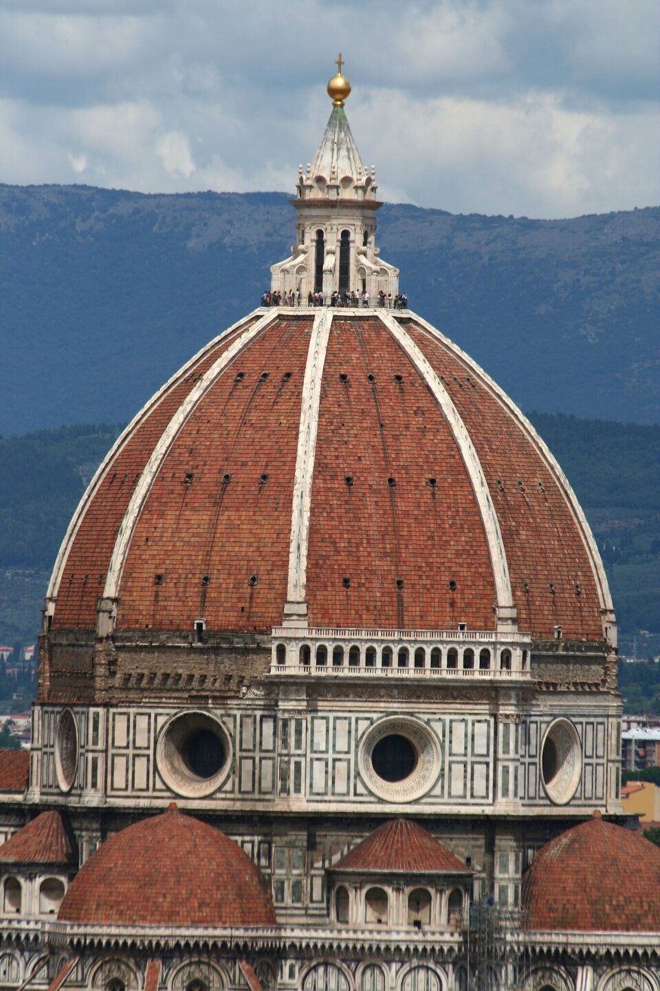 Kolorowa fotografia przedstawiająca monumentalną kopułę katedry o czerwonej, ceglanej dachówce, podzieloną białymi kamiennymi żebrami. U podstawy kopuły widoczny jest tambur z dużymi, okrągłymi oknami i dekoracyjnymi arkadami. Na szczycie wznosi się smukła latarnia zakończona złotą kulą i krzyżem; wokół jej podstawy stoją drobne sylwetki ludzi na tarasie widokowym. W tle rozciąga się górzysty krajobraz pod pochmurnym niebem.