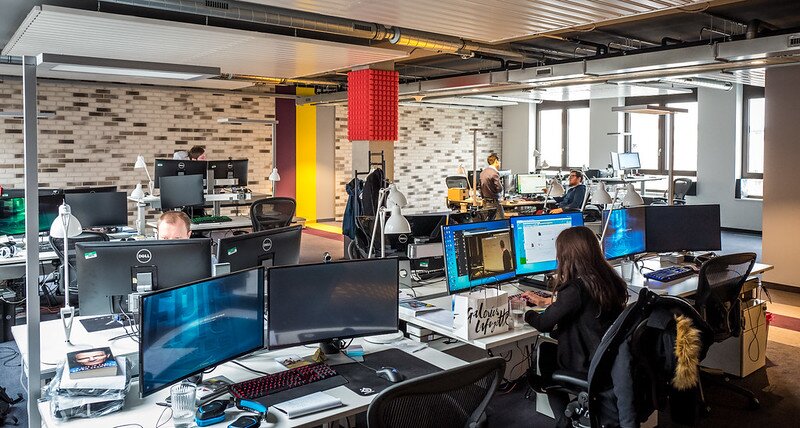 Zdjęcie przedstawia wnętrze biura firmy z branży informatycznej. W pomieszczeniu znajduje się wiele mniejsc do pracy. Są biurka poustawiane obok siebie, na nich są komputery, klawiatutry, myszki, przy nich stoją krzesła biurowe. Na większości biurek stoją po dwa komputery obok siebie, przy niektórych z nich siedzą pracownicy.