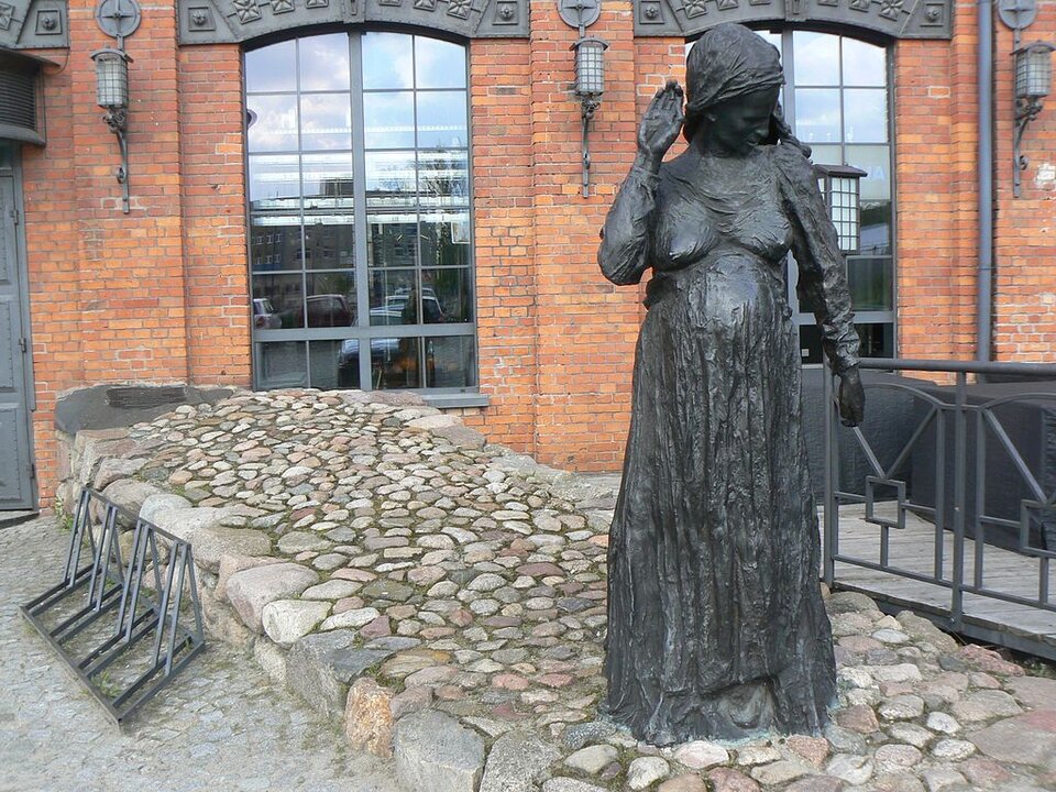 Zdjęcie przedstawia pomnik kobiety w długiej sukni i z chustą na głowie. Kobieta stoi, ma uniesioną prawą rękę. Pomnik stoi na tle budynku z czerwonej cegły i z wysokimi oknami.