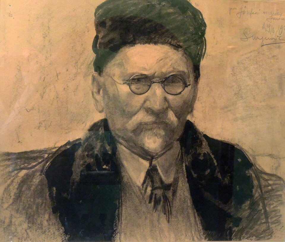 Obraz przedstawia portret starszego mężczyzny w okularach, ubranego w ciemne odzienie z futrzanym kołnierzem oraz zieloną czapkę. Jego twarz jest surowa, pomarszczona, a spojrzenie poważne. Artysta użył techniki rysunku z elementami malarskimi, węglem i akwarelą lub pastelami, co nadaje pracy ekspresyjny charakter. Tło jest oszczędne, co sprawia, że cała uwaga skupia się na twarzy i wyrazie postaci. W prawym górnym rogu widoczne są odręczne napisy i podpis, sugerujące dedykację lub autograf autora.