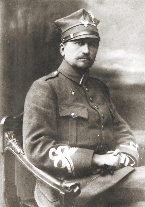 Ilustracja przedstawia żołnierza. Jest to mężczyzna w średnim wieku z wąsem, ma zamyśloną twarz. Siedzi na krześle ubrany jest w mundur, na głowie ma wojskową czapkę z orłem białym.