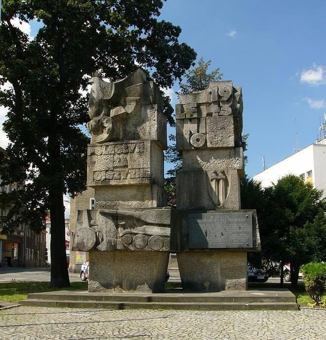 Zdjęcie przedstawia betonowy pomnik. Jest on zbudowany z dwóch kolumn, na których umieszczone są między innymi napisy: poległym za ojczyznę, Lenino 1943. Widać także płaskorzeźby ukazujące: orła, czołg, dwa pionowo ustawione miecze obok siebie.